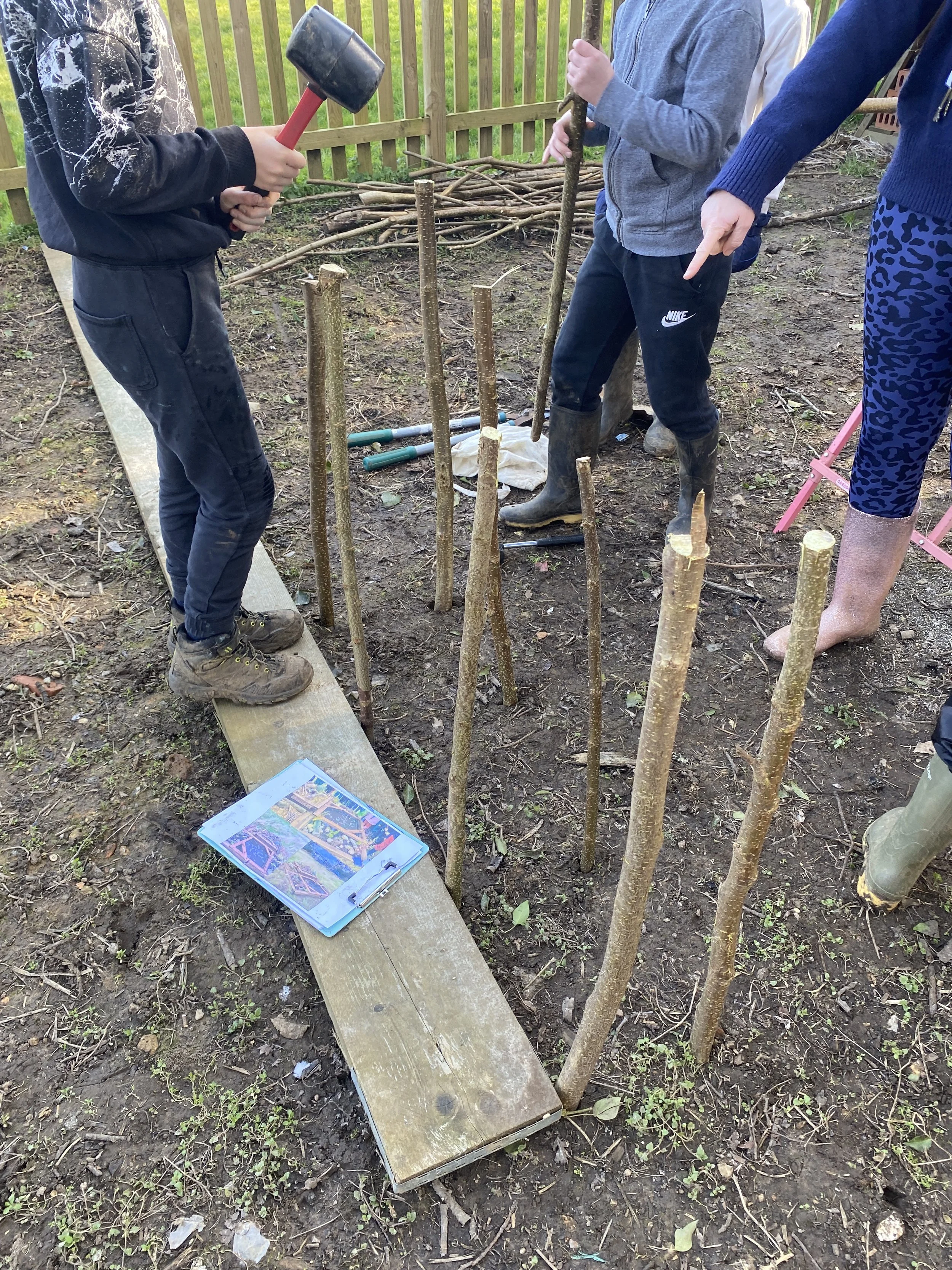 Children planting trees in a backyard, with saplings, a wooden plank, and a guidebook on the ground.