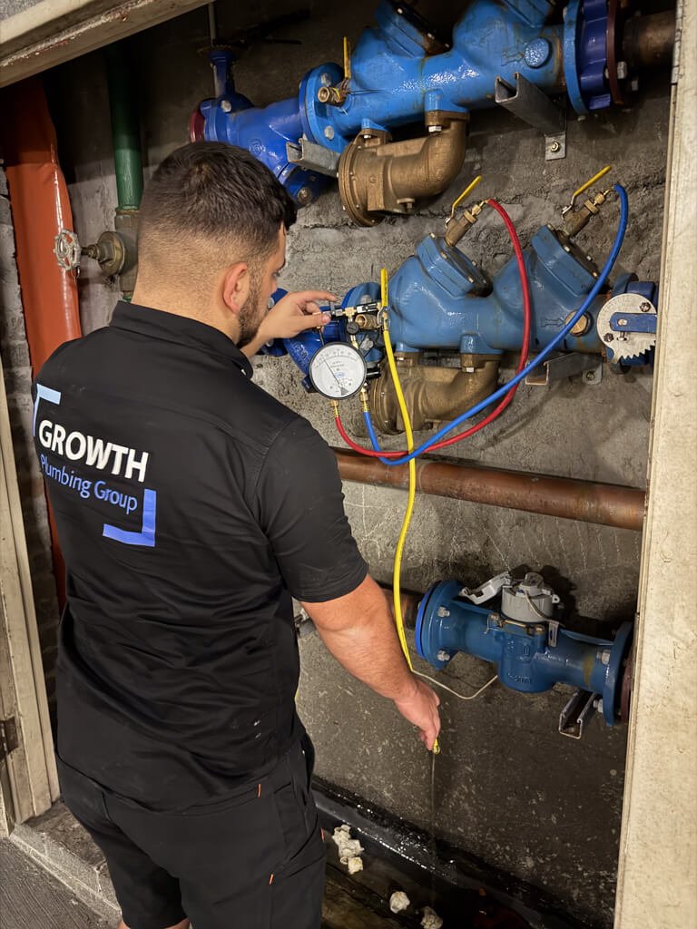 Plumber working on water meter and pipes, wearing a black shirt with the logo 'GROWTH Plumbing Group', inside a utility closet.