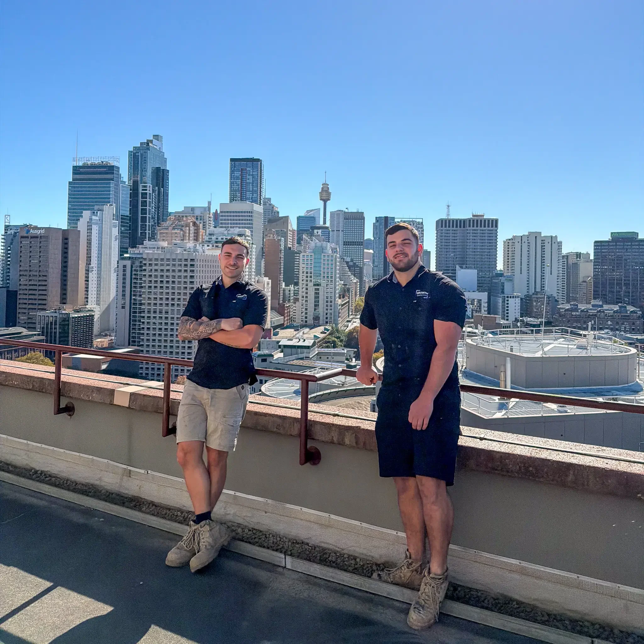 Two men standing on a rooftop with a city skyline in the background, both people are dressed in black shirts and wearing tactical boots.