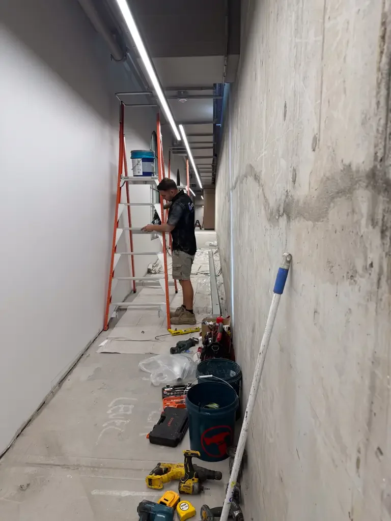 A worker standing on an orange scaffold painting an interior wall with a paint roller, with tools and supplies scattered on the floor.