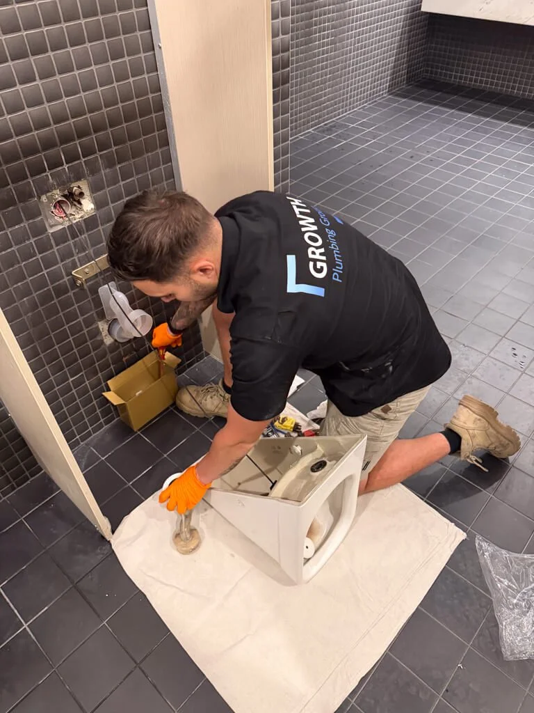 Plumber repairing or replacing a toilet in a bathroom with black tiled floors and walls.