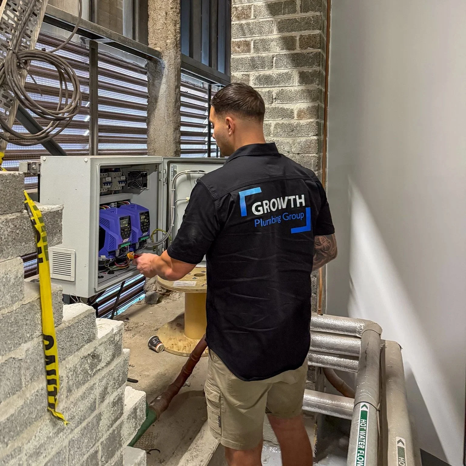 A technician wearing a black shirt with 'Growth Plumbing Group' working on an electrical panel in a building with brick and concrete walls, and pipes running along the floor.