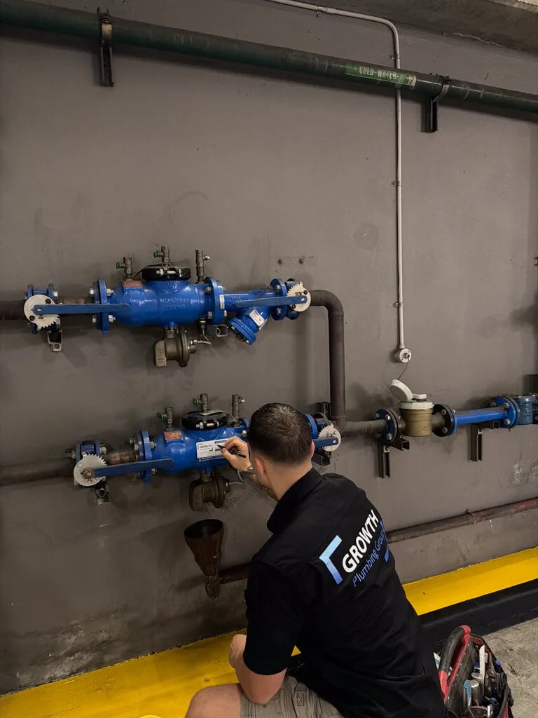 A man working on plumbing pipes in an industrial setting, wearing a black shirt with the logo 'GROWTH Plumbing Group'.