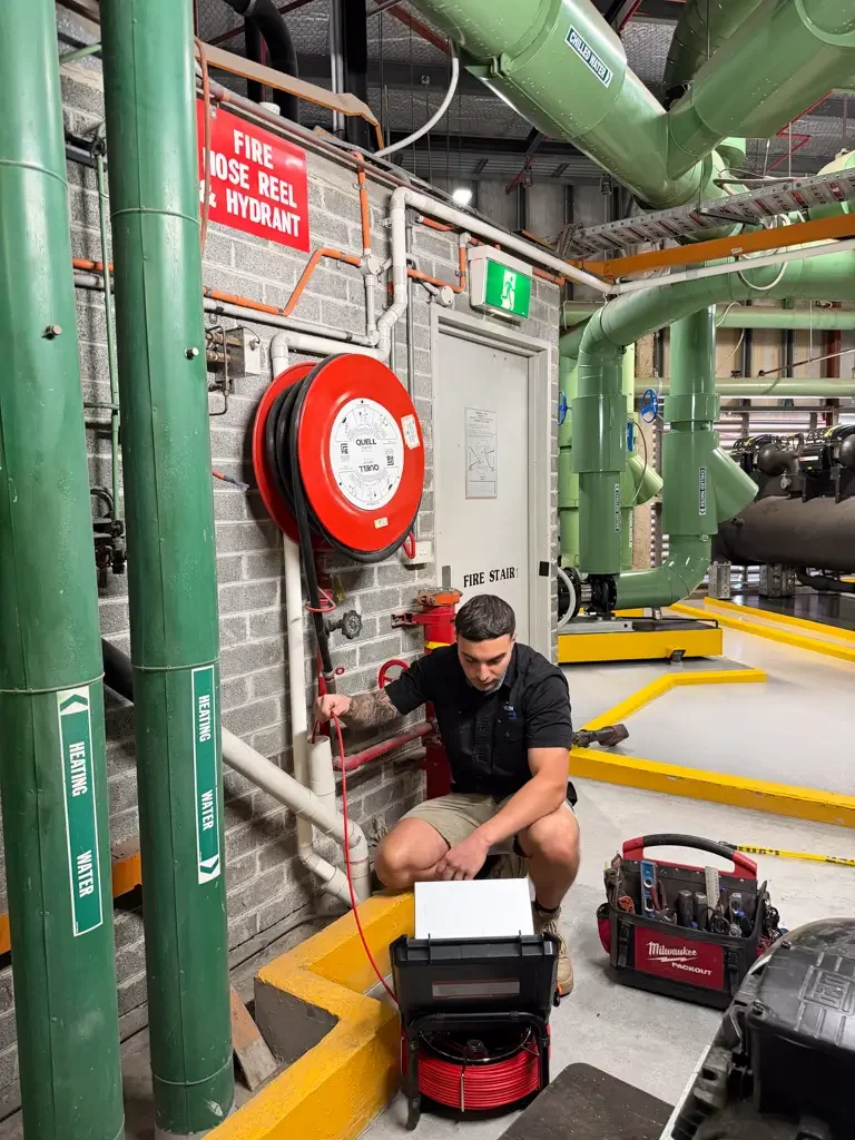 A man is working in an industrial setting with green pipes, a red fire hose reel, and safety signs, including a fire hydrant sign and a fire exit sign. He is squatting on a concrete floor next to a toolbox and a red reel of cable.
