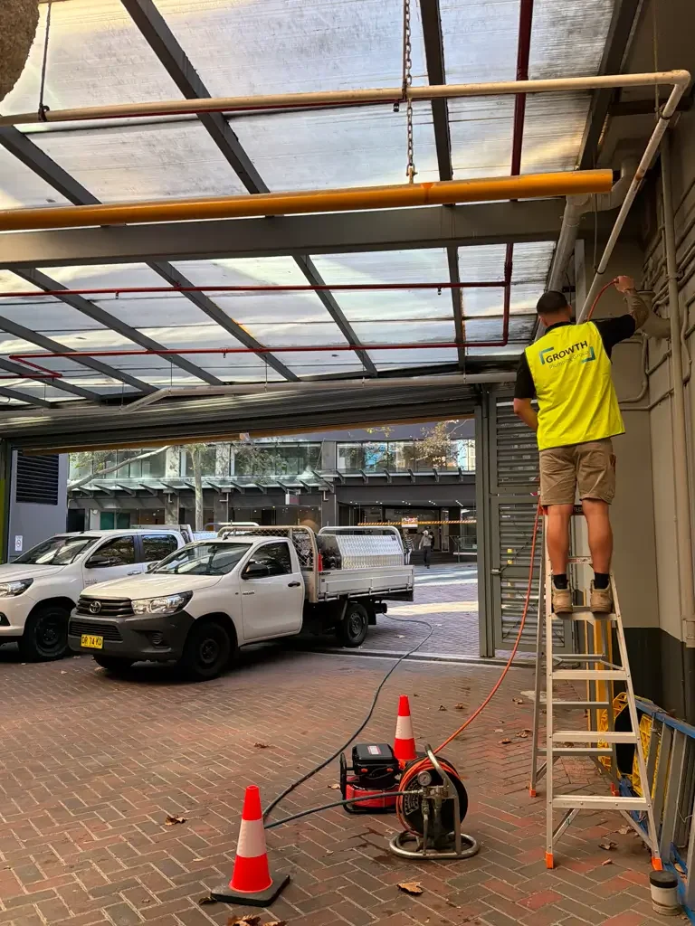A worker on a step ladder using a tool under a roof structure to inspect or repair pipes or electrical wiring.