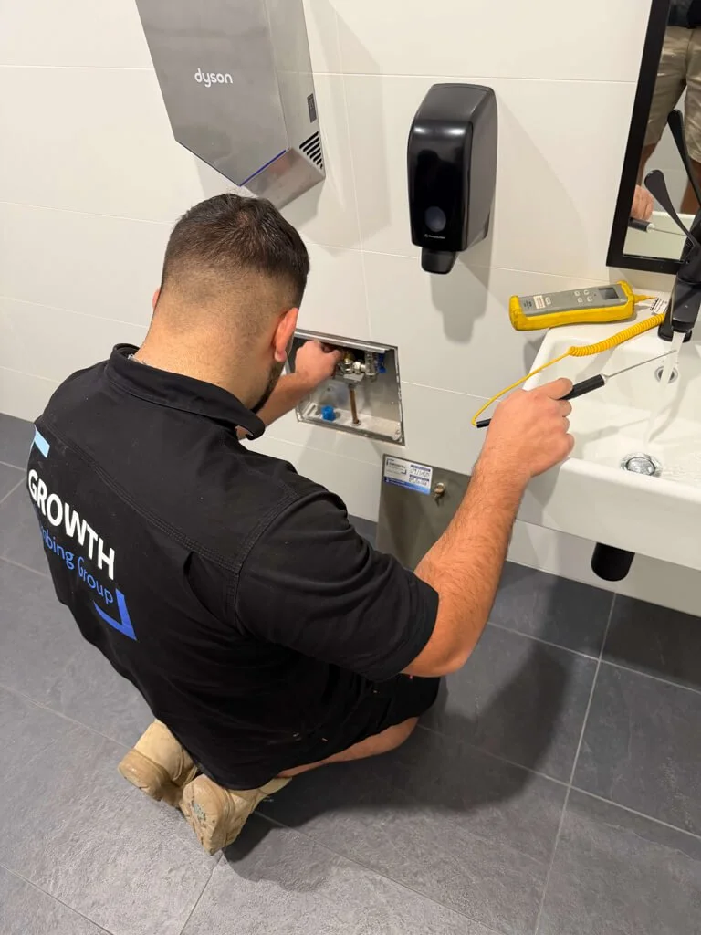 Technician kneeling on the floor, working on plumbing under a bathroom sink. The technician is wearing a black shirt with a company's logo, and tools are on the sink counter, including a multimeter and a screwdriver.