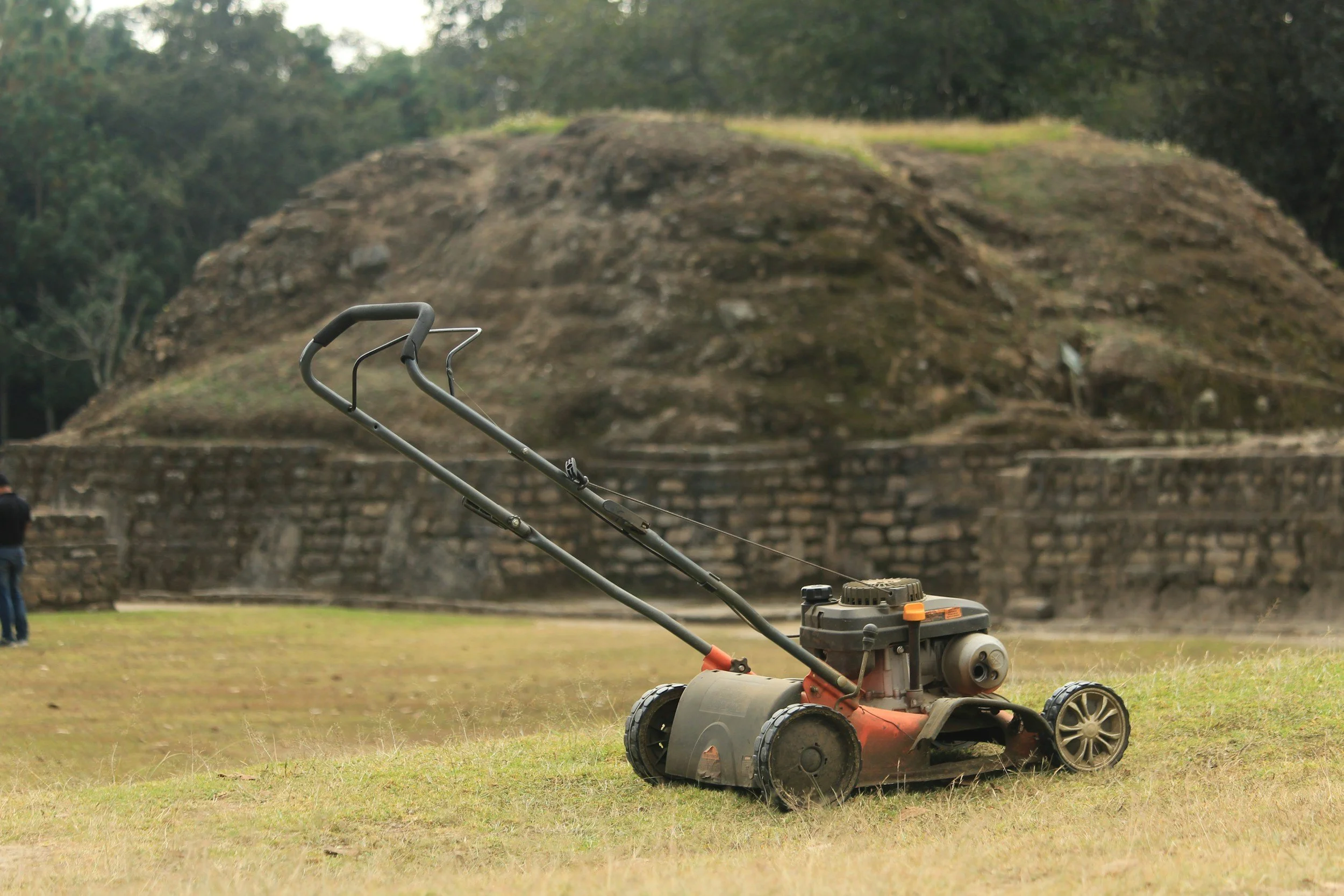 Lawnmower sitting on grass with ancient ruins and lush green trees in the background.