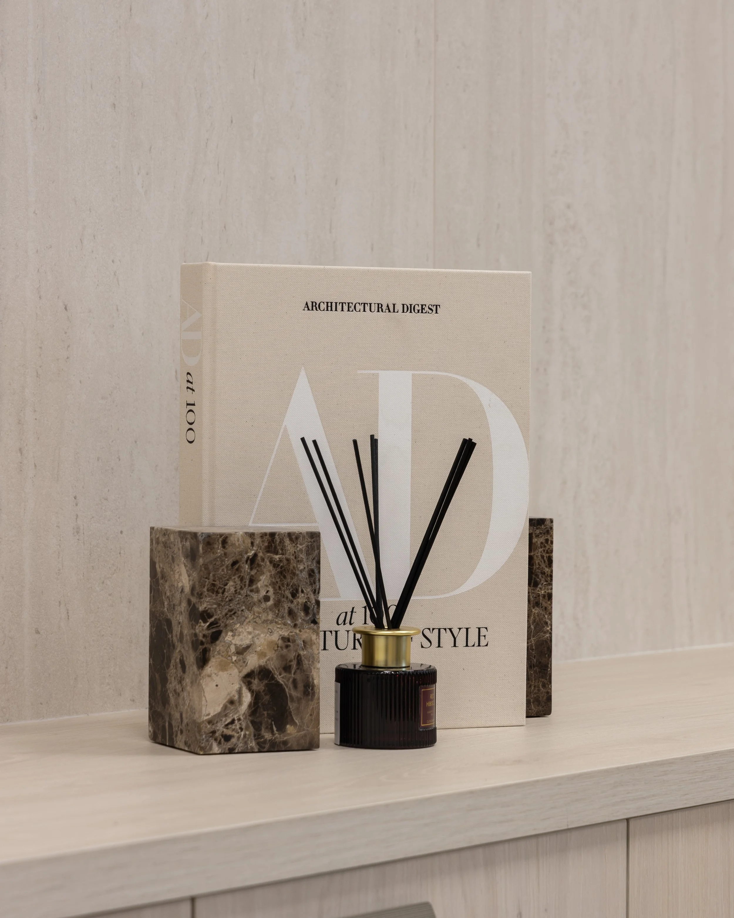 A marble vase with dark and light brown patterns, a black candle jar with a gold lid, a large beige book titled 'ARCHITECTURAL DIGEST' with white and black text, all arranged on a white shelf against a beige wall.