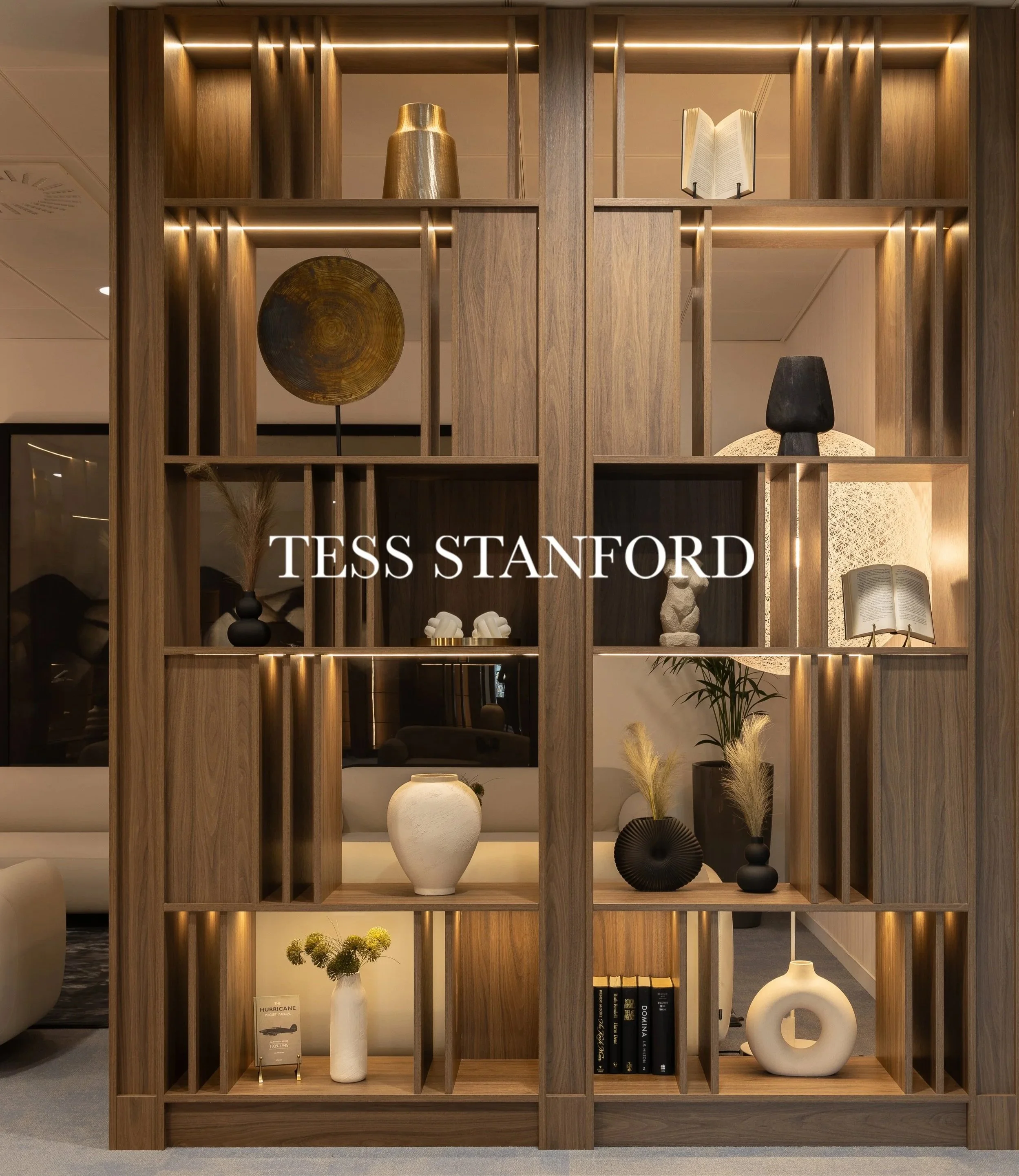 A wooden room divider shelf displaying decorative items including a gold vase, an open book on a stand, a wooden platter, a black head sculpture, white and black vases, a potted plant, and a small plant in a white vase with a label that reads 'HURRICANE.' The shelf has built-in lighting and is part of a modern interior design.