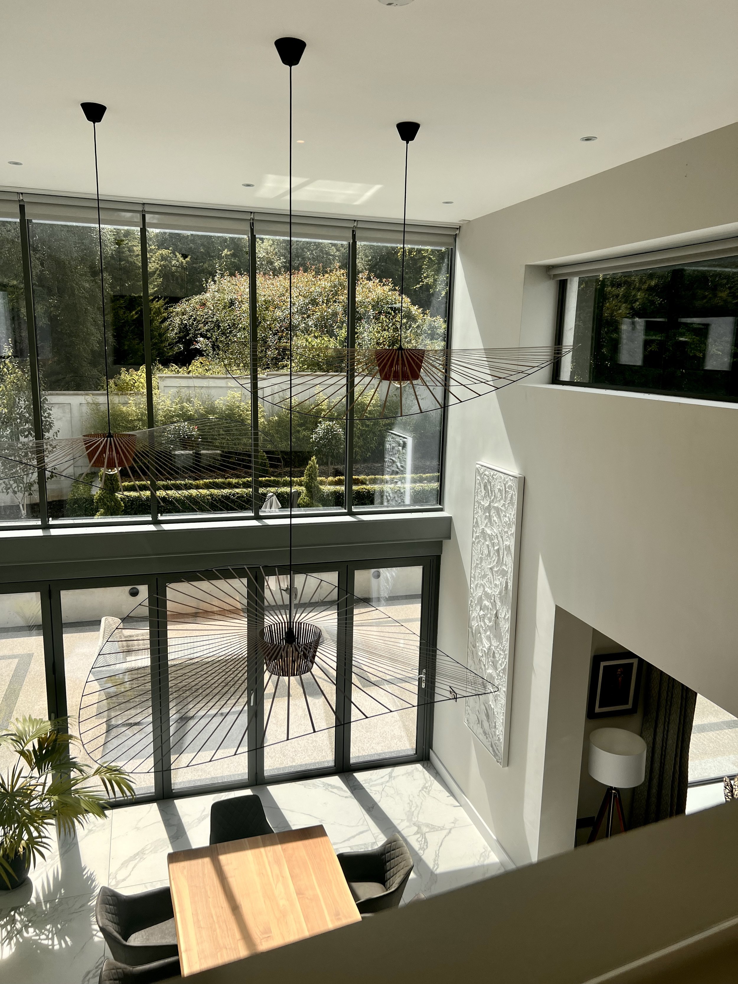 A modern interior living space viewed from an upper level, featuring a wooden dining table with black chairs, large glass windows overlooking a lush garden with trees, and contemporary hanging light fixtures.