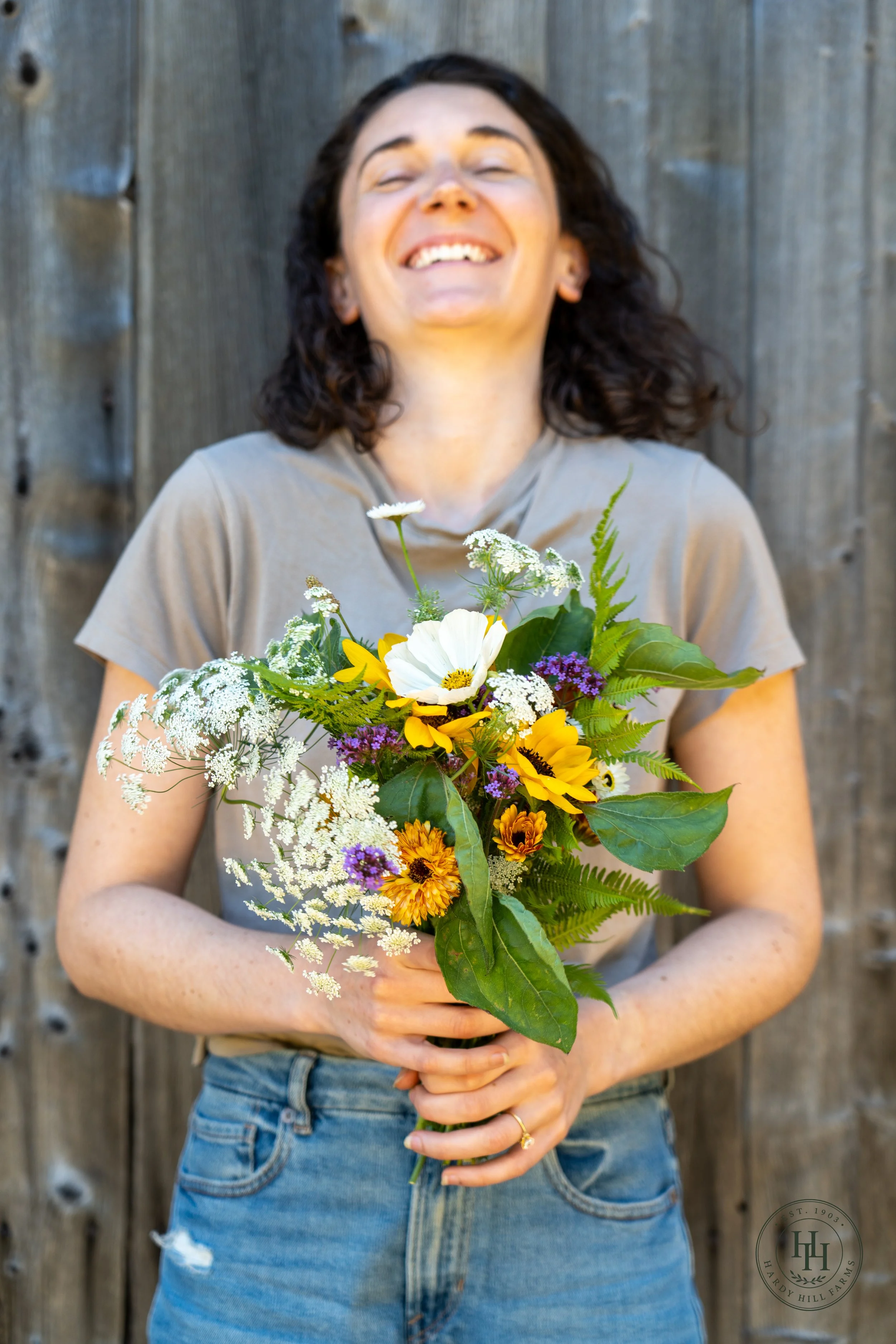 A woman with dark curly hair smiling with eyes closed, holding a vibrant bouquet of various flowers in front of a wooden fence.