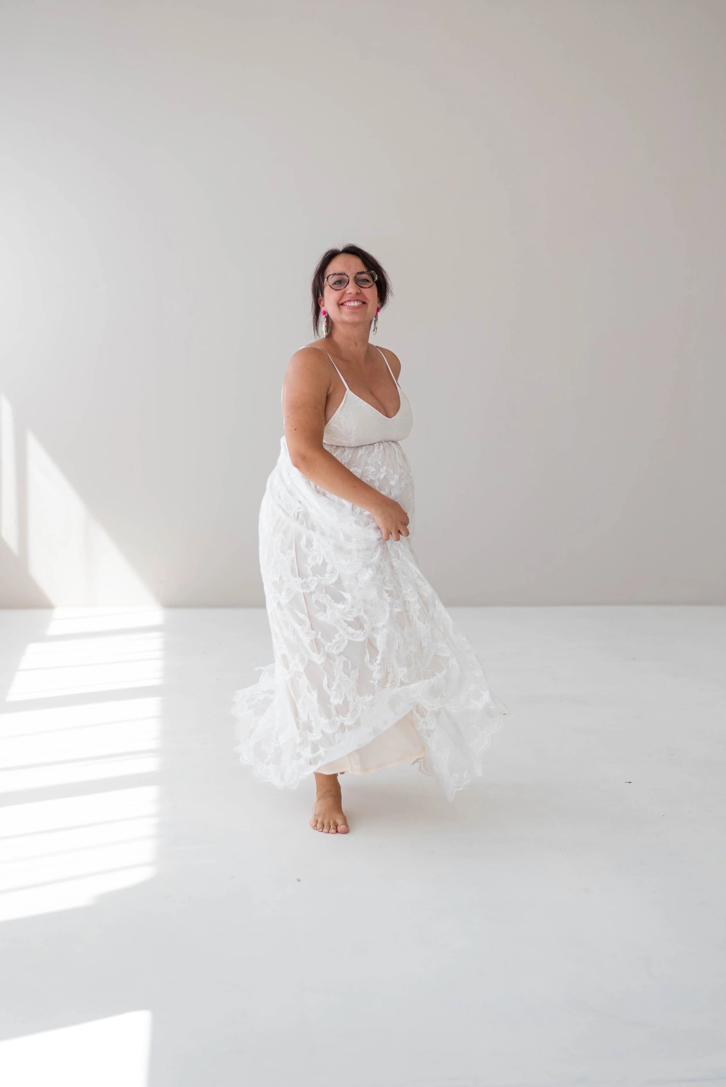 A woman with glasses, earrings, and a white dress is smiling and walking barefoot in a bright, minimalist white room.