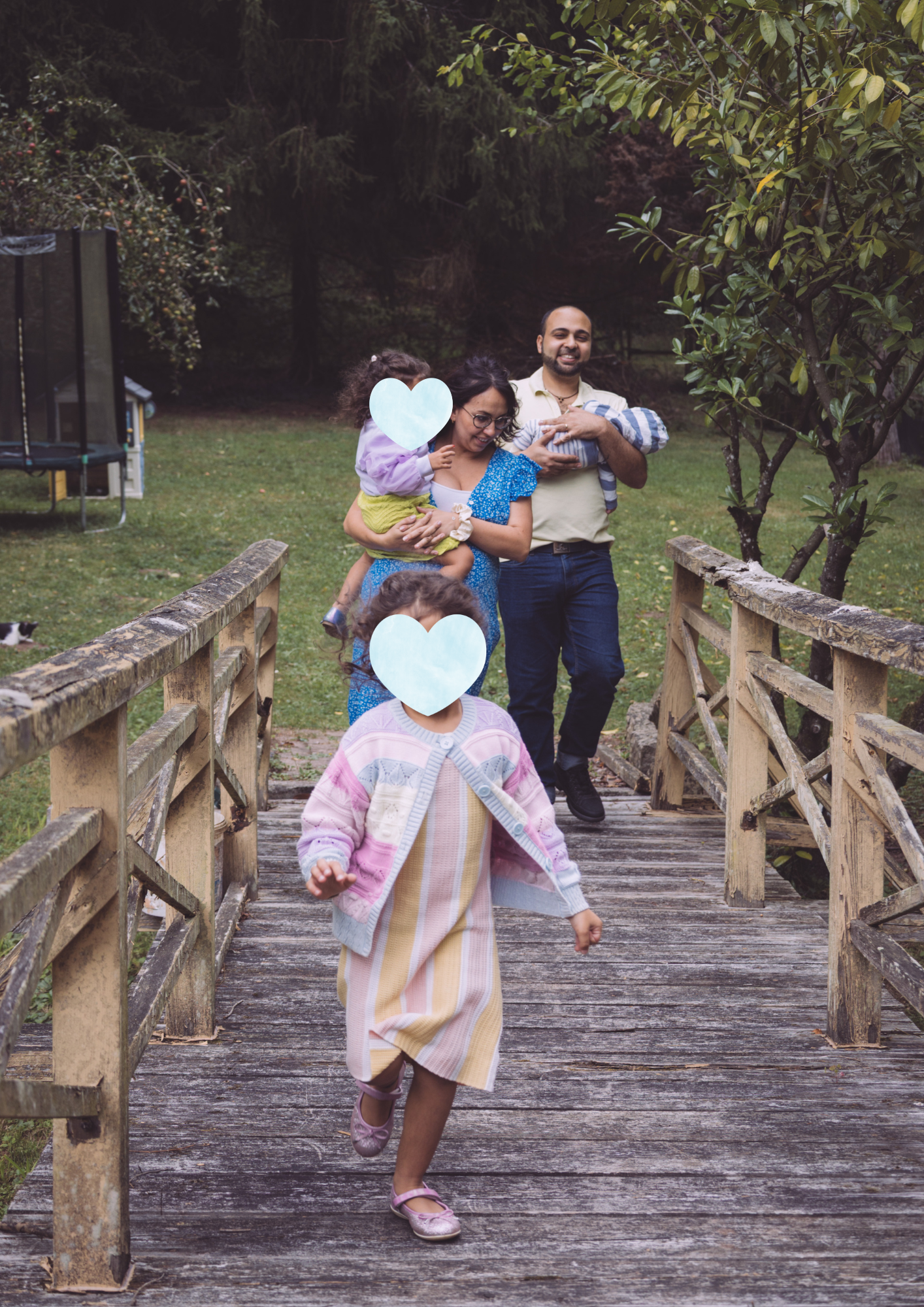 Family walking on a wooden bridge outdoors in a park or garden, with children and adults smiling, and trees in the background.