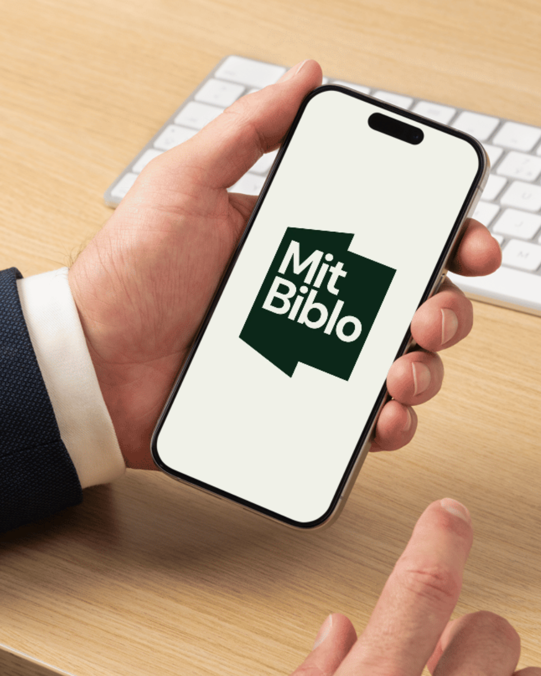 En person holder en smartphone med en app kaldet 'Mit Biblo' på skærmen, med et tastatur i baggrunden.