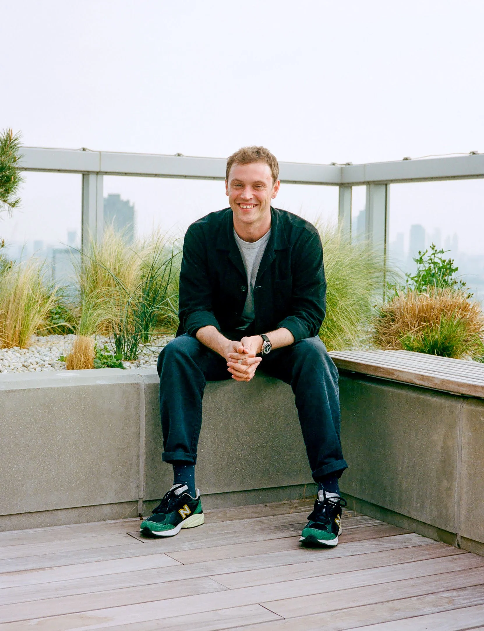 A young man smiling and sitting on a concrete ledge on a rooftop balcony with plants and a city skyline in the background.