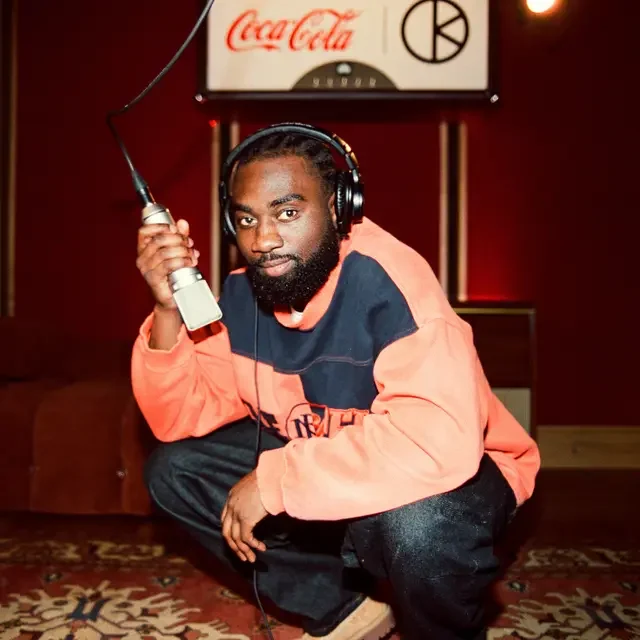 A man with a beard wearing headphones, a colorful jacket, and black pants crouches on a rug in front of a Coca-Cola sign, holding a microphone.