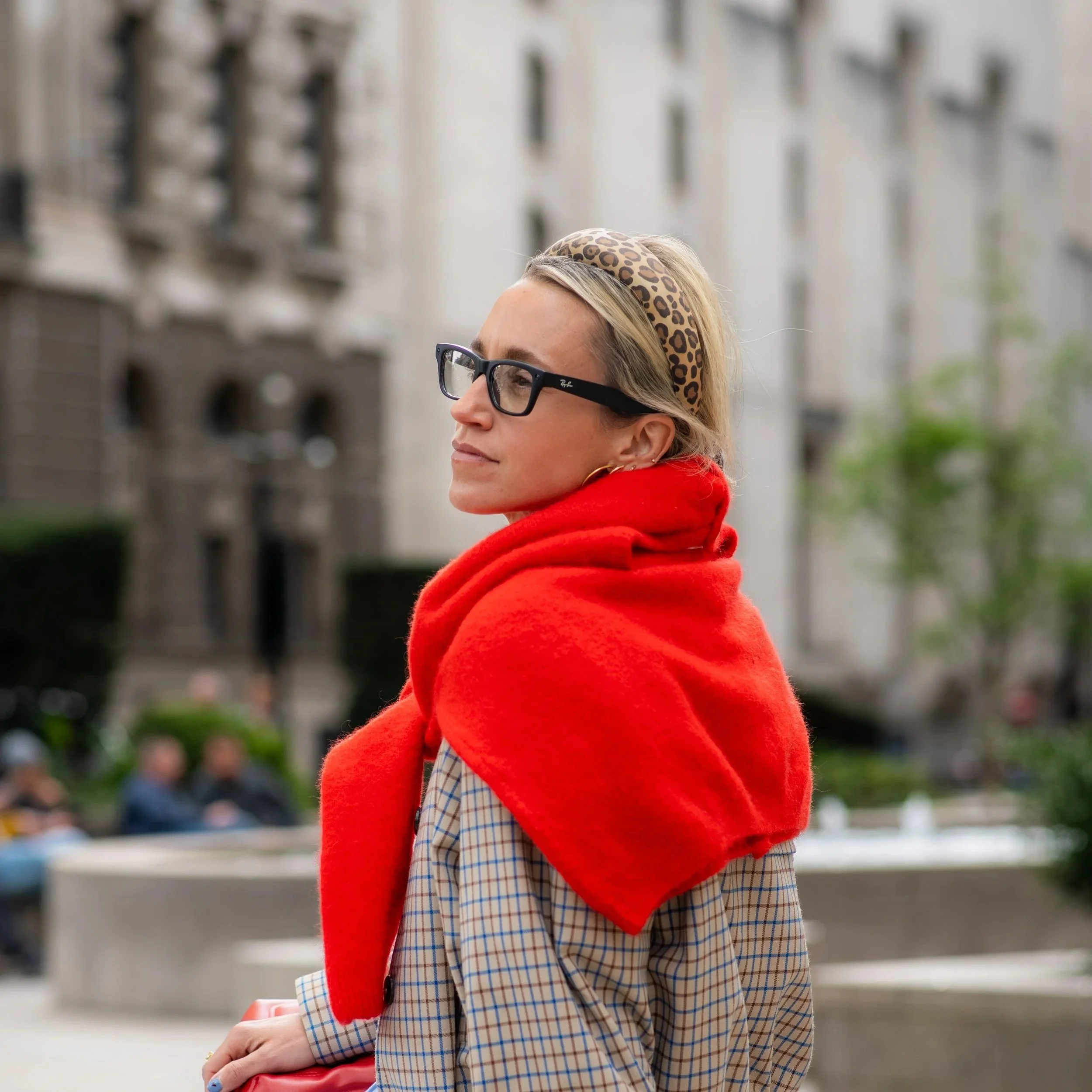 A woman with blonde hair, wearing glasses, a leopard print headband, a red scarf, and a plaid blazer, standing outdoors with blurred buildings and trees in the background.