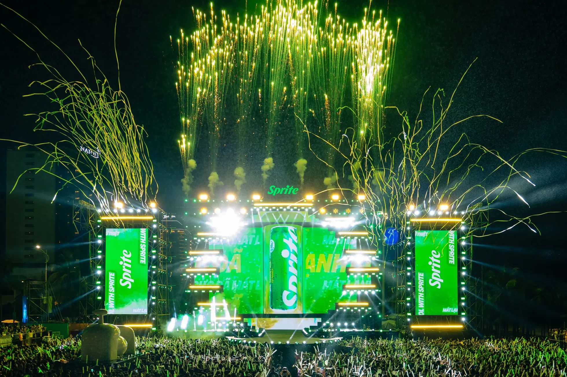 Fireworks display over a concert stage with green and yellow lighting, advertising banners for Sprite, and a large crowd watching.