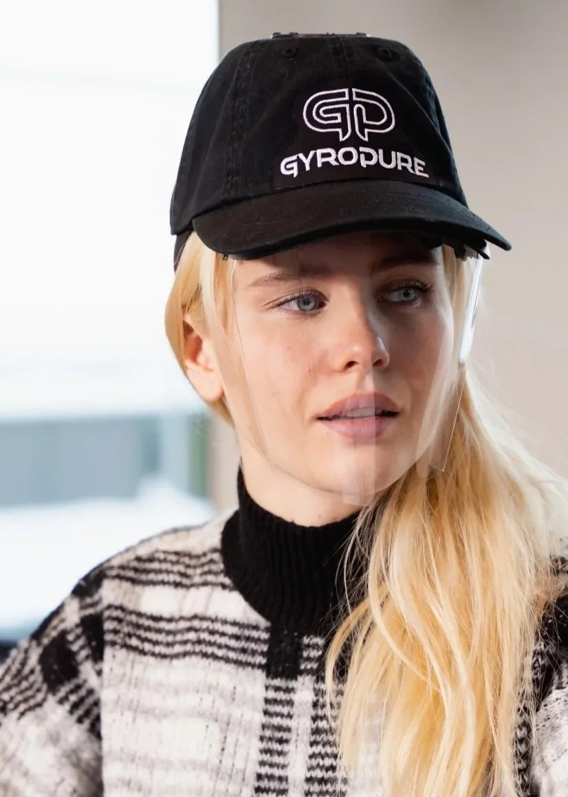 A young woman wearing a black cap with the GYPODURE logo and a patterned sweater, near a window.