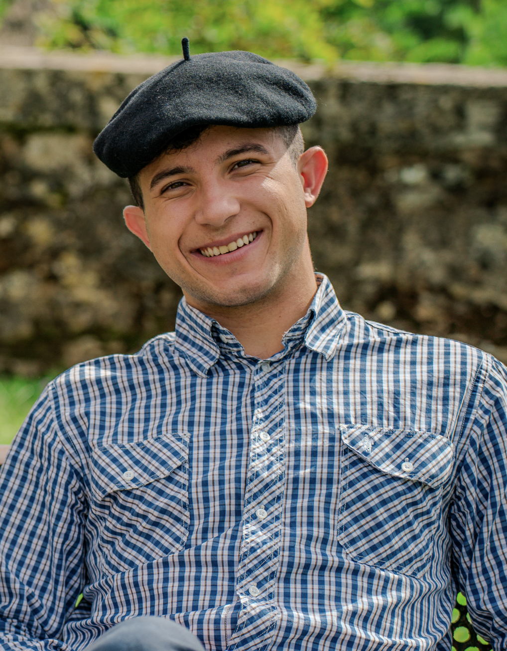 Jeune homme souriant portant un béret noir et une chemise à carreaux bleus, avec un arrière-plan naturel.