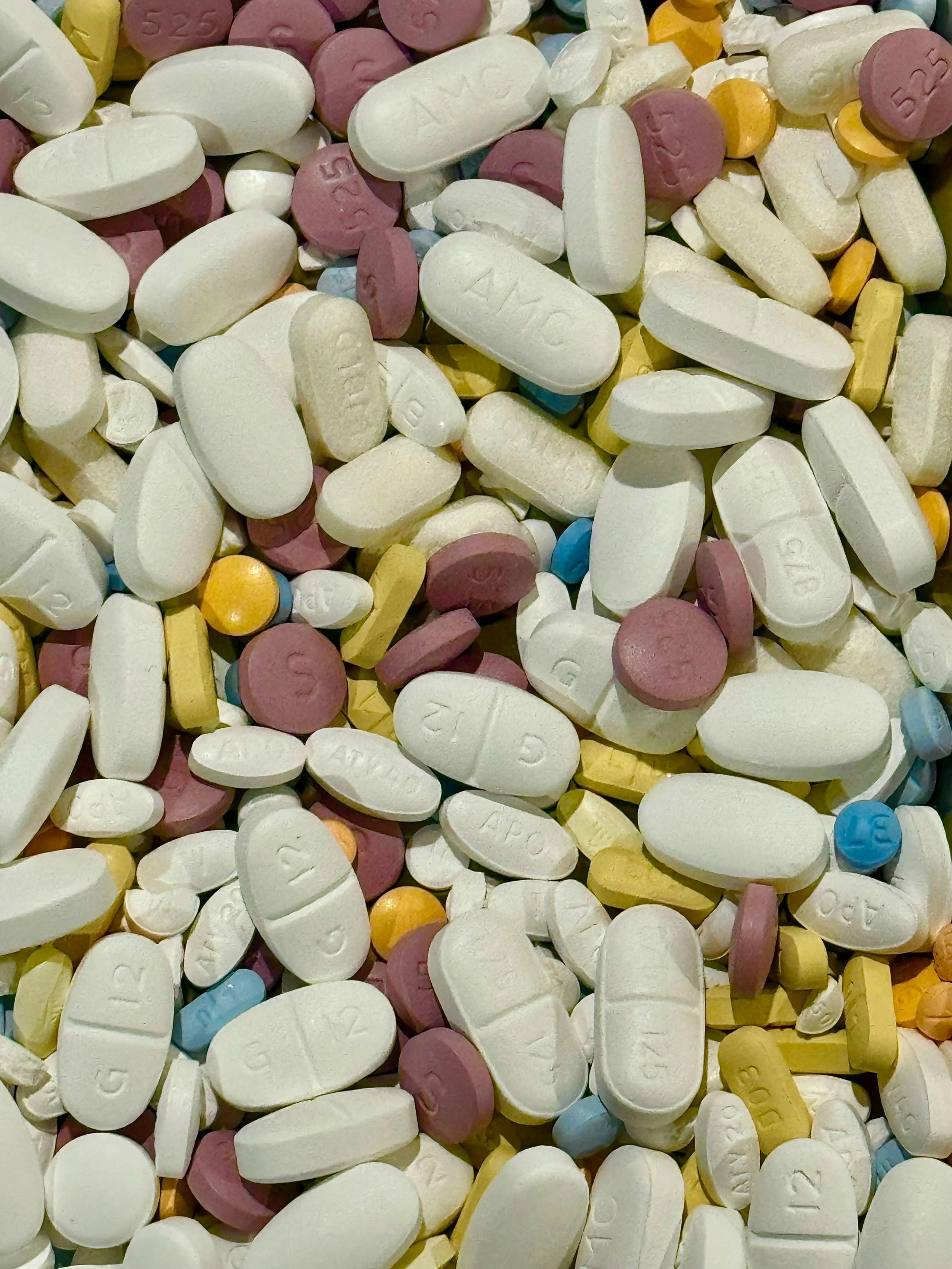 A close-up of numerous assorted pills, including oval and round shapes, in colors such as white, pink, yellow, and blue, with various imprints and markings.