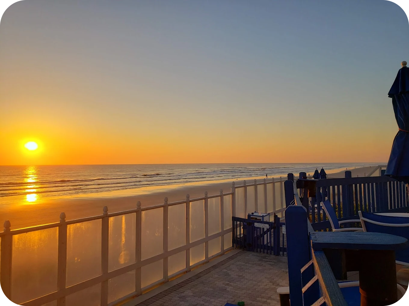 Sunset over the ocean viewed from a beachfront patio with blue chairs and umbrellas, and a white fence.
