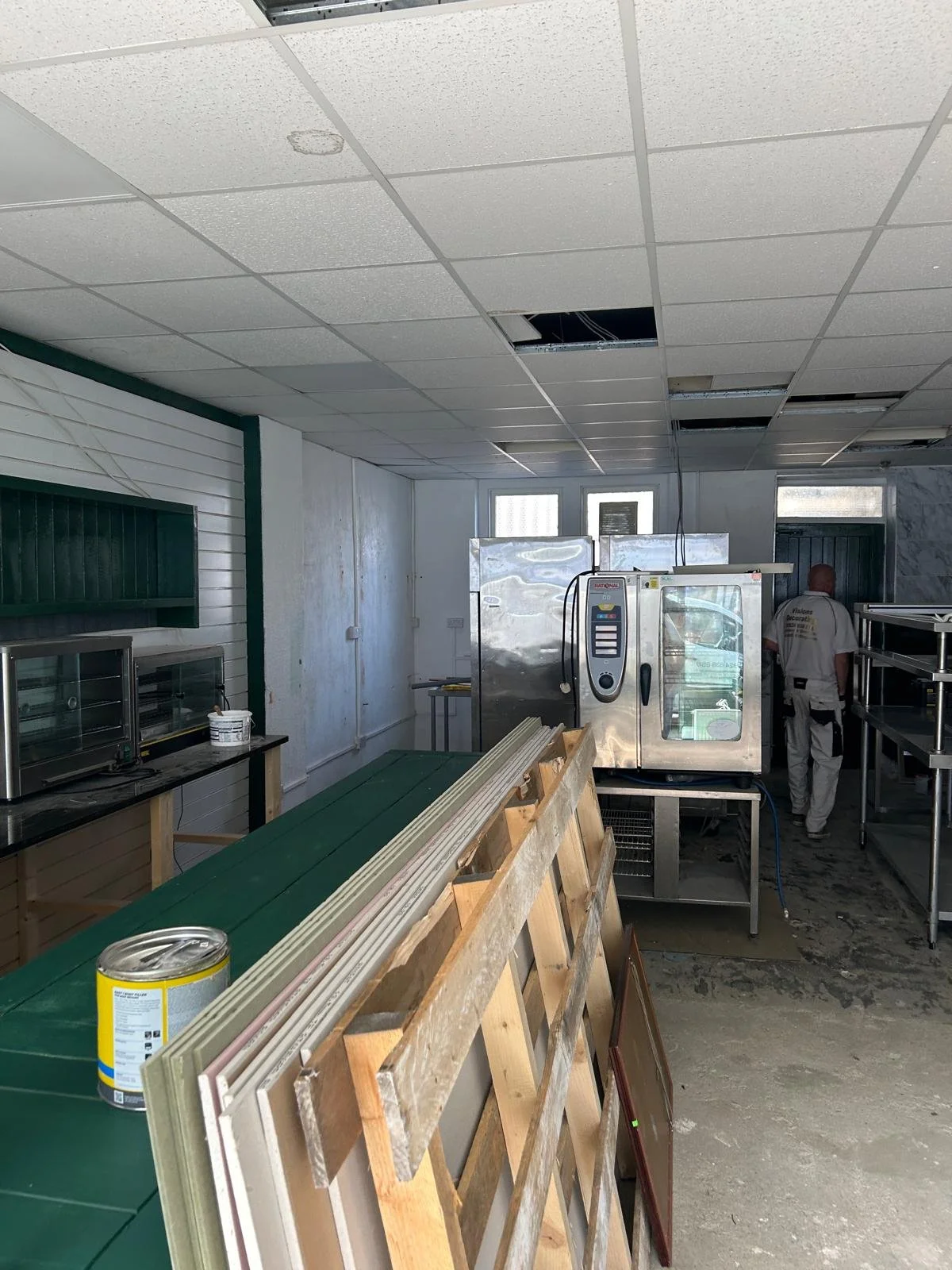 Construction site or renovation of a commercial kitchen, with ceiling tiles removed, a worker, and equipment like an oven, shelving, and construction materials.