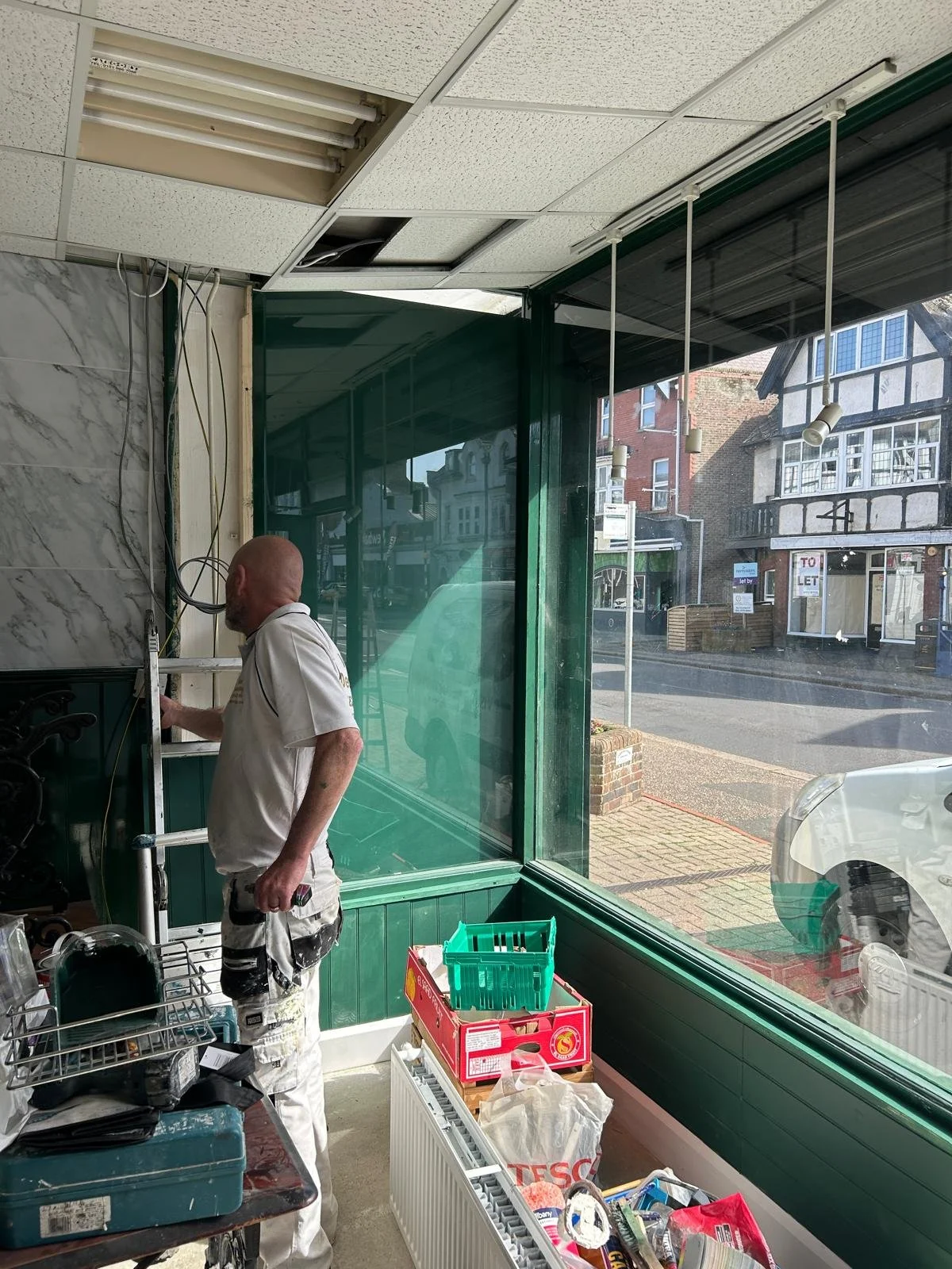 A man with a bald head is working inside a store, standing near a counter with boxes of supplies, and looking at some equipment or items. The store has large front windows showing a street scene outside with buildings, parked cars, and a 'To Let' sign.