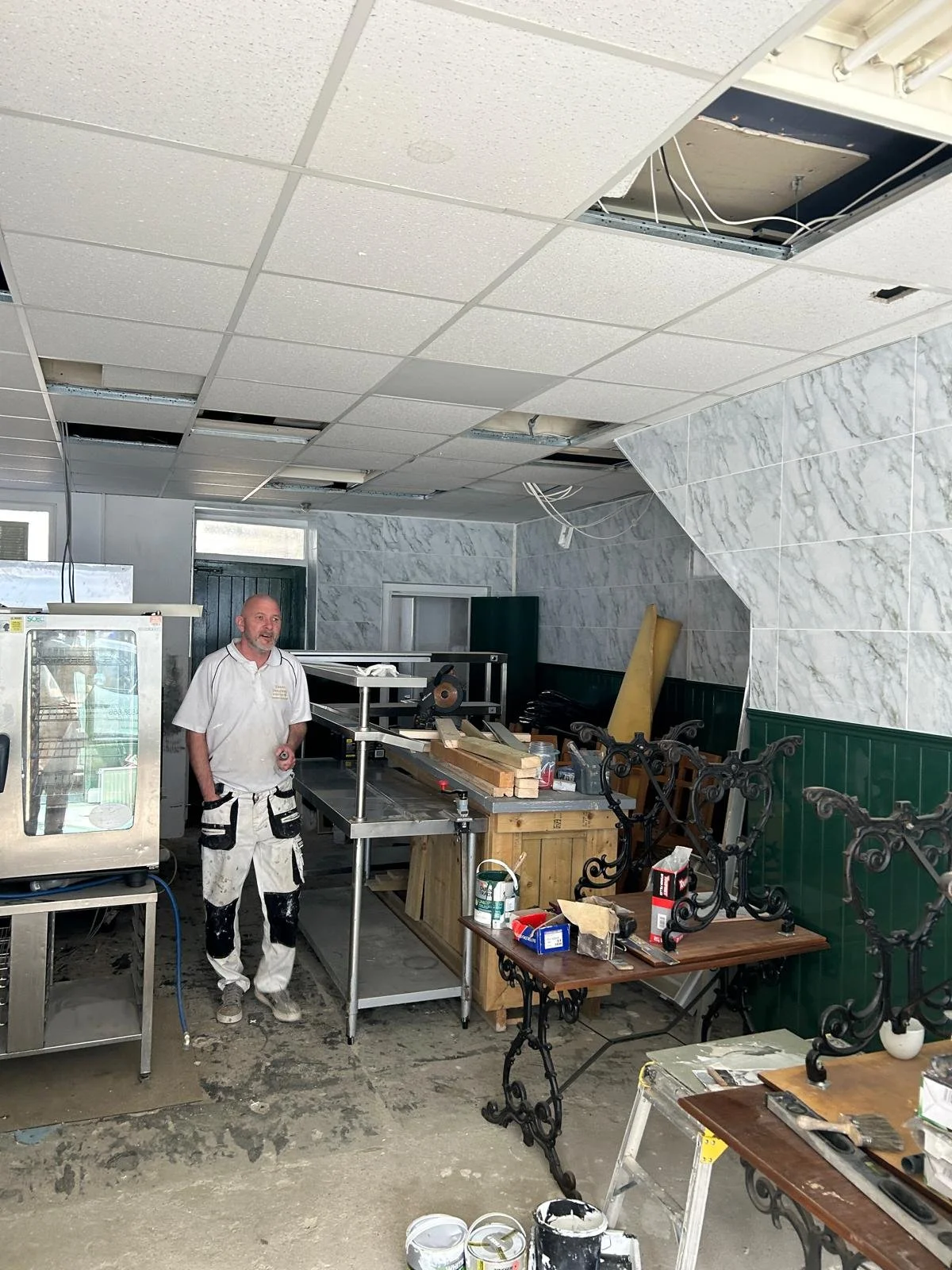 A man in work overalls standing in a restaurant under renovation, with a partially removed ceiling and construction tools and materials around.