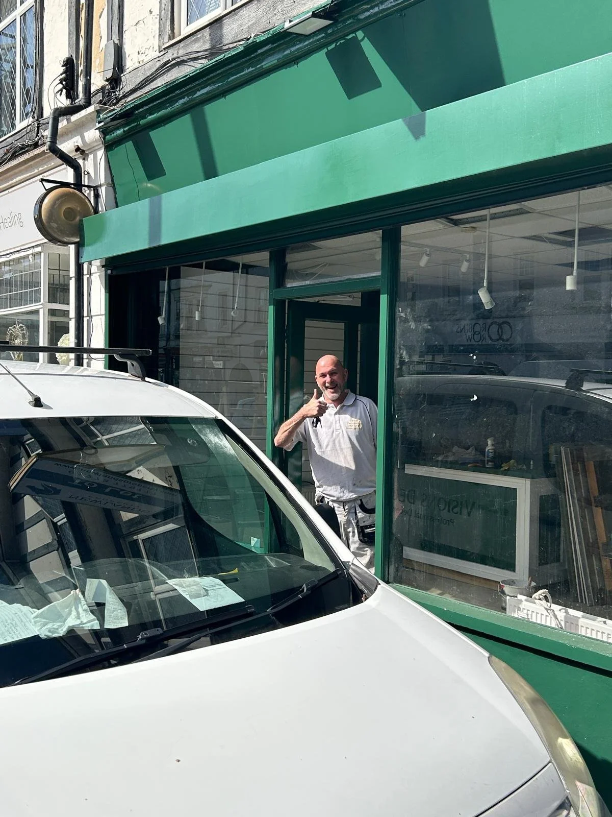 A man standing inside a storefront, smiling and giving a thumbs-up gesture, visible through a large glass window. A white vehicle is parked in front of the store.