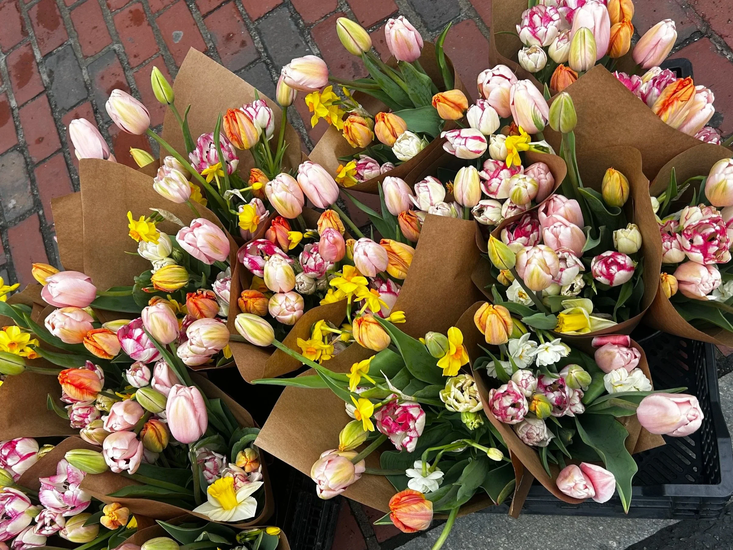 Bundles of colorful tulips and daffodils wrapped in brown paper are displayed on a brick sidewalk.