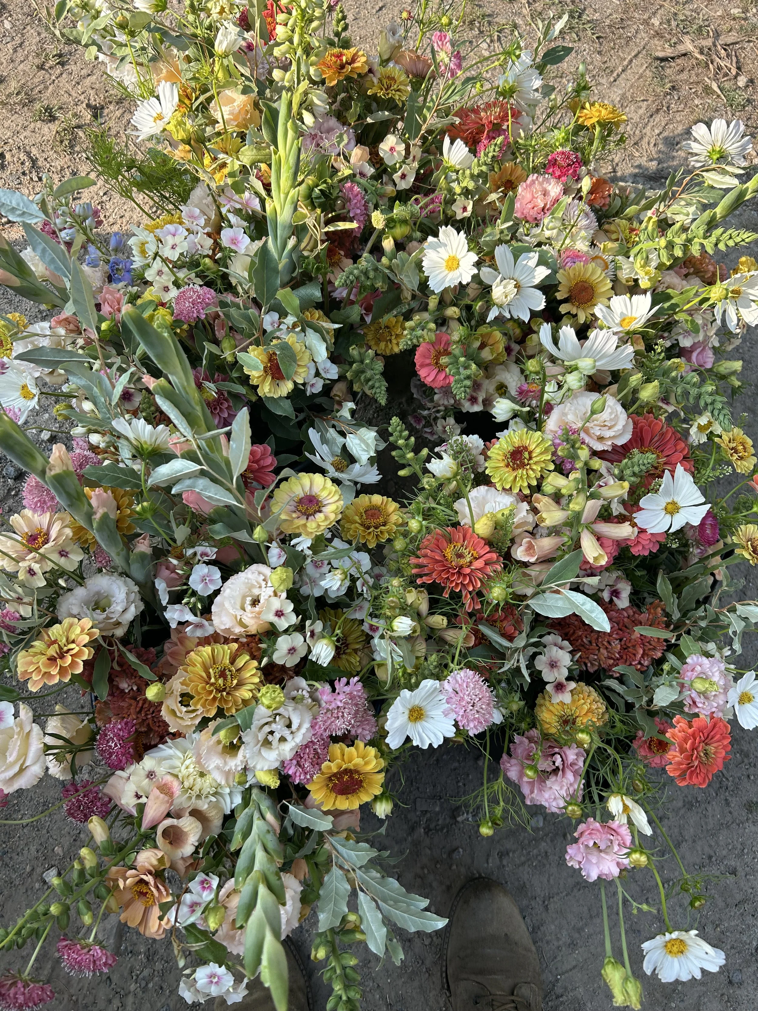 A colorful assortment of flowers including daisies, carnations, and other blossoms placed on the ground with a person's shoes visible at the bottom of the image.