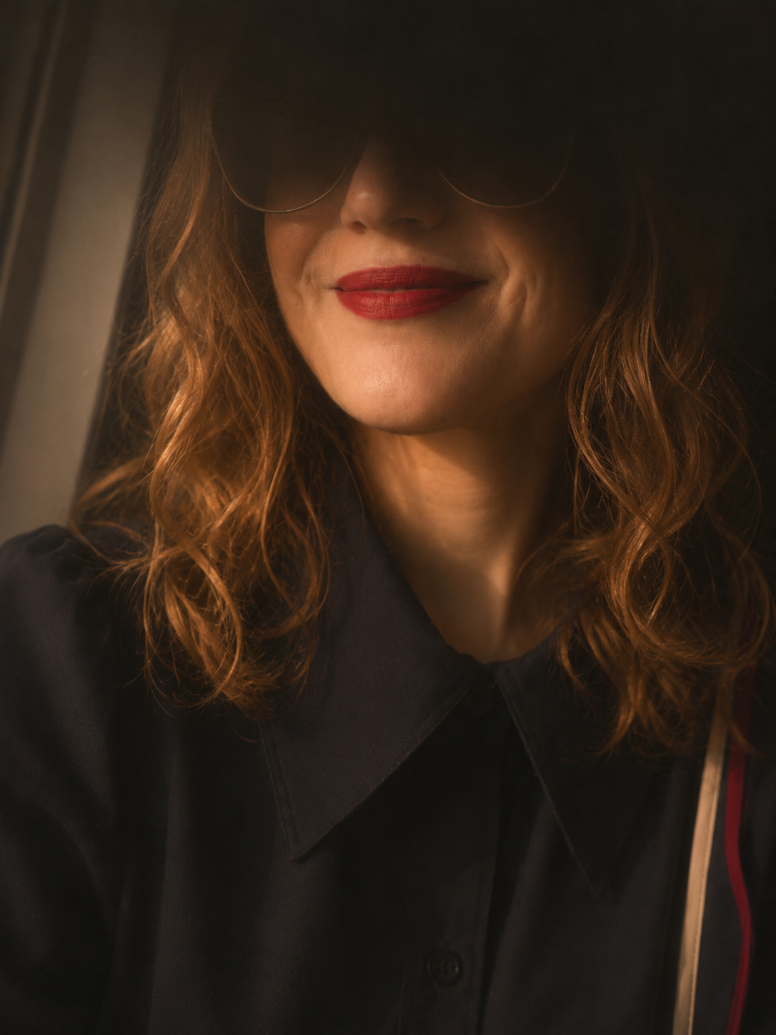 Close-up of a woman with brown hair, wearing round sunglasses, a dark shirt with a collar, and red lipstick. She is partially hidden in a dark environment.