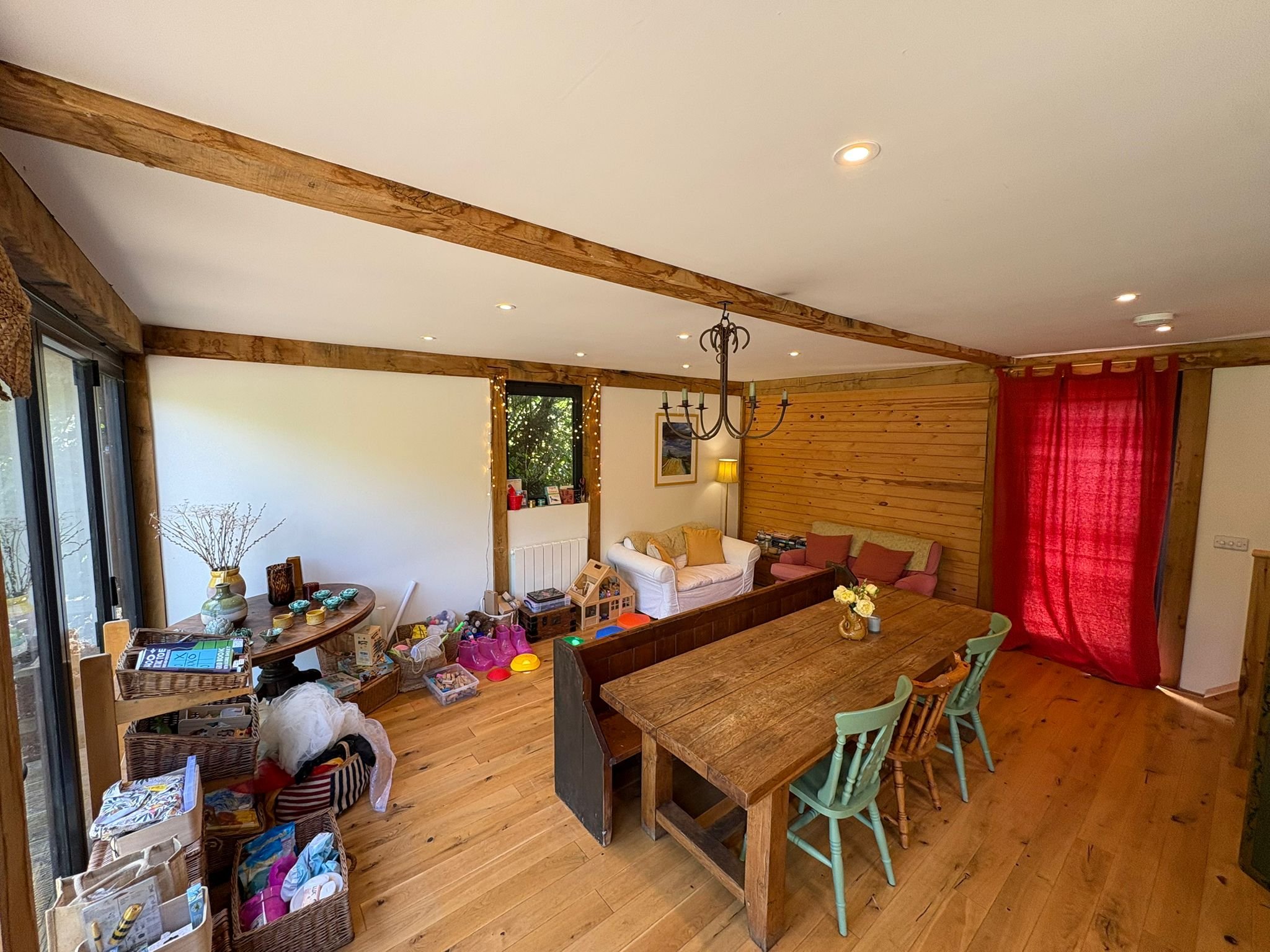 Living room with wooden beams, a chandelier, red curtain, and furniture, with toys and clutter on the floor.