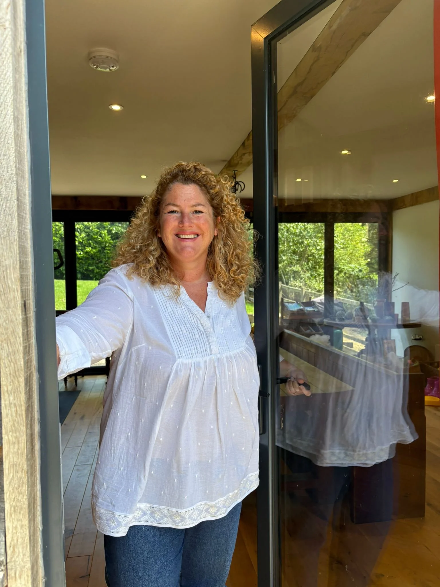 A woman with curly blonde hair smiling and looking at the camera, standing in a room with large windows and wooden accents, as viewed from a doorway with the door open.