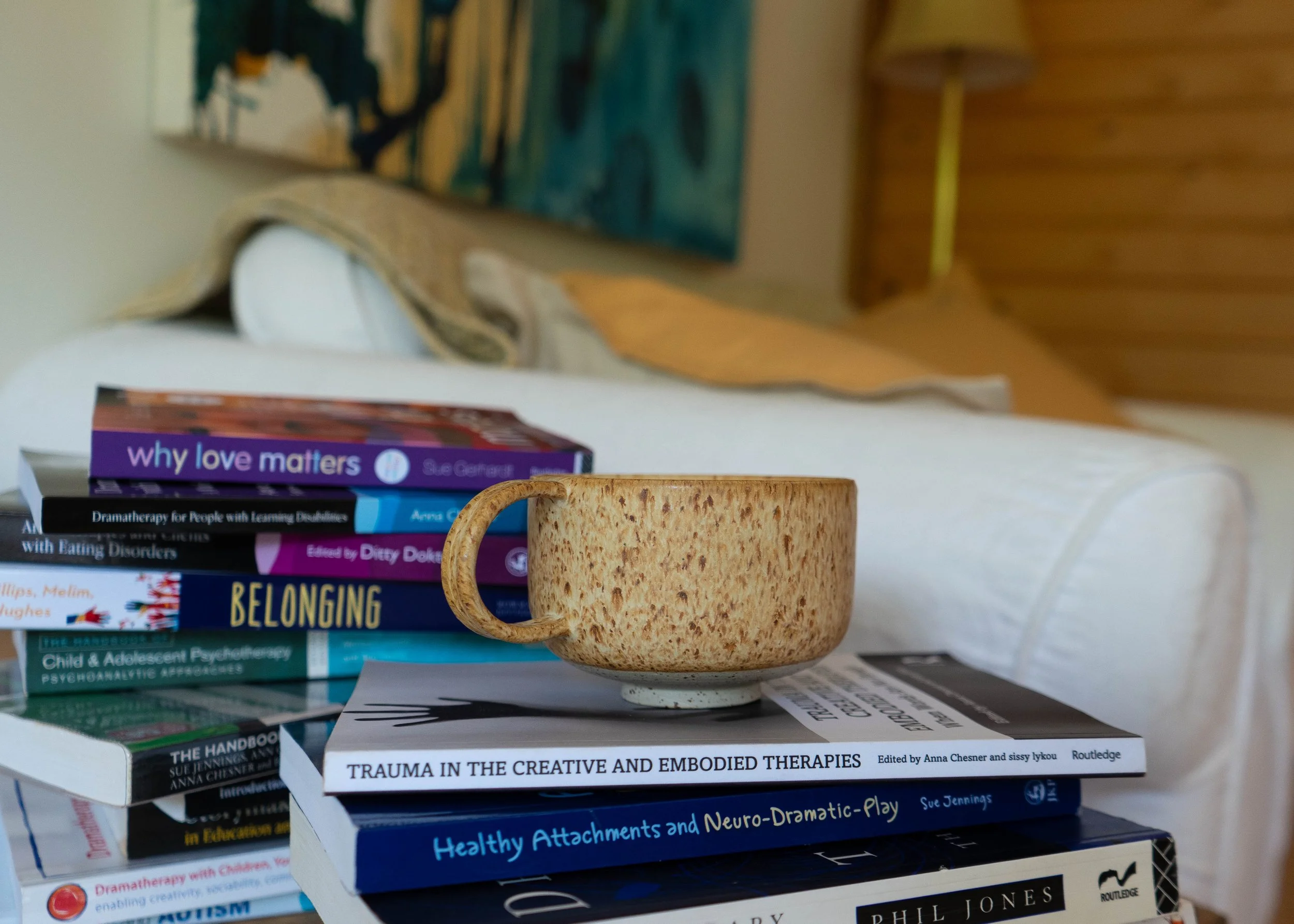 A stack of books on a table, with a ceramic mug in the foreground. The books include titles related to therapy, childhood, trauma, attachment, neuro-drama, and psychology.