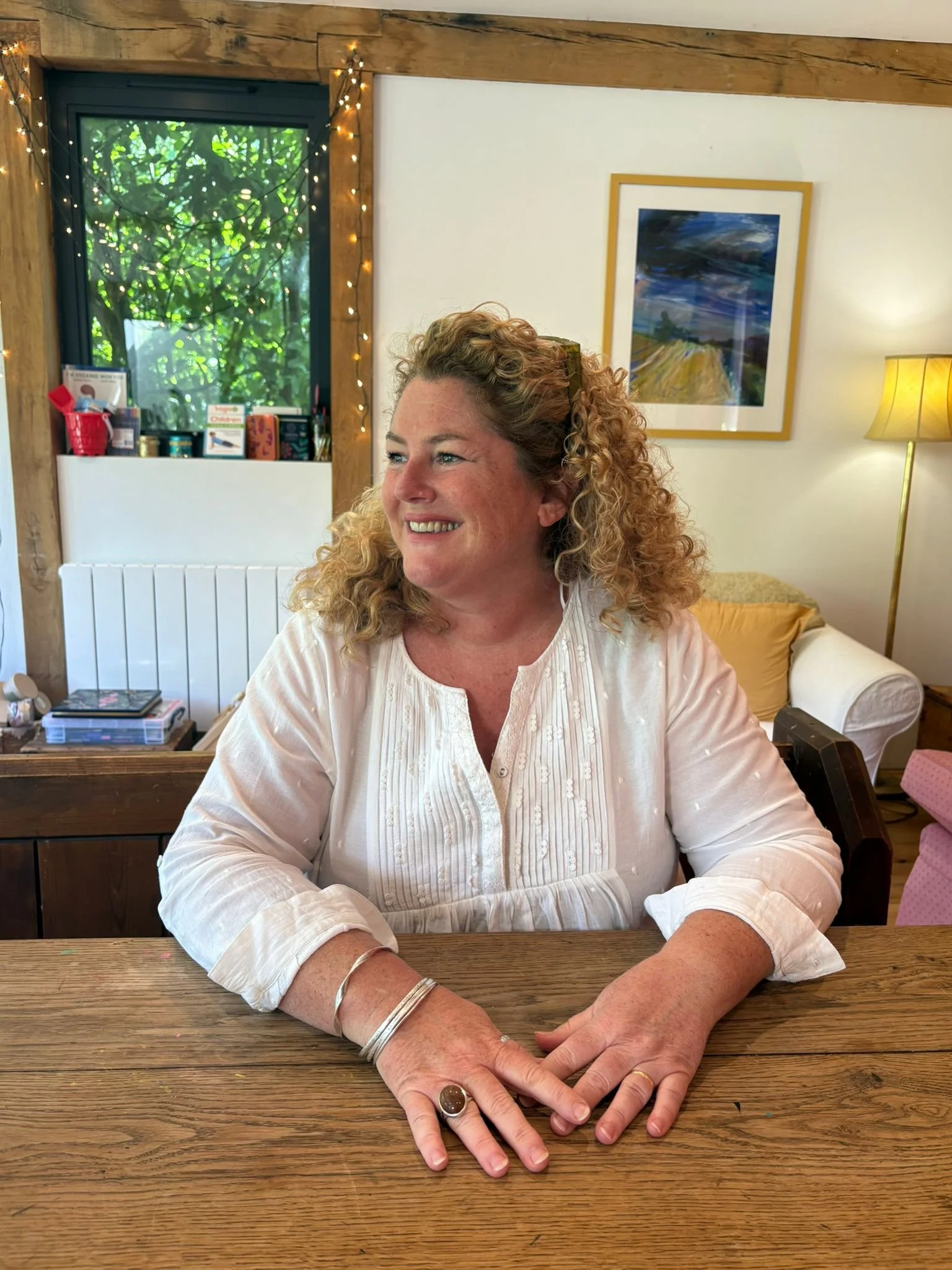 A woman with curly hair and fair skin sitting at a wooden table, smiling and looking to her left. She is wearing a white blouse with embroidery, and jewelry including a large ring and silver bracelets. Behind her is a white wall with a framed landscape painting, a yellow lamp, and a window showing greenery outside. The room is decorated with string lights and various items on a shelf.
