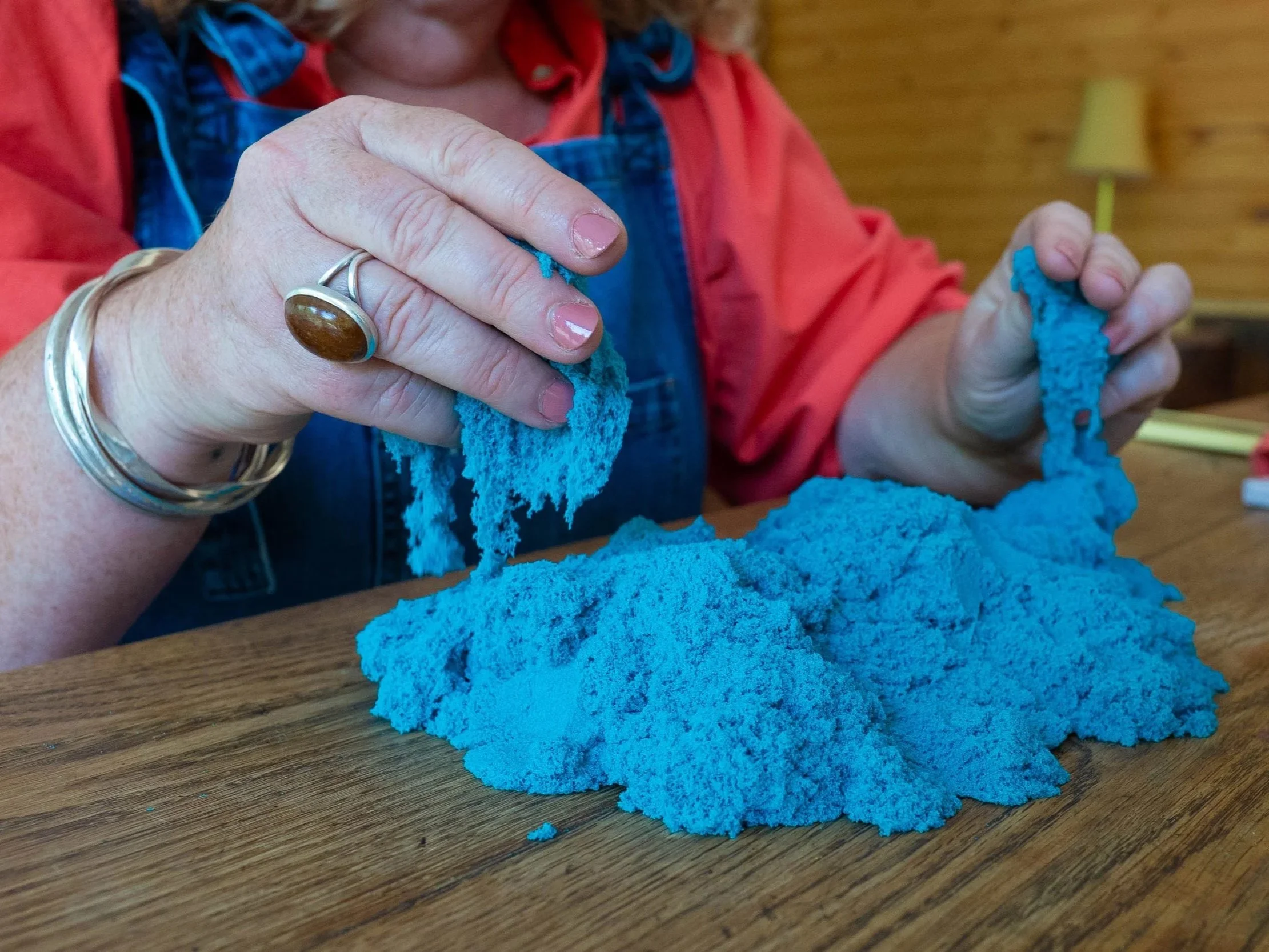 Close-up of a person's hands holding a piece of bright blue yarn, with a pile of similar yarn on a wooden table. The person is wearing several silver rings and a bracelet, and has painted fingernails. The background shows a blurred indoor setting, possibly a cozy room.