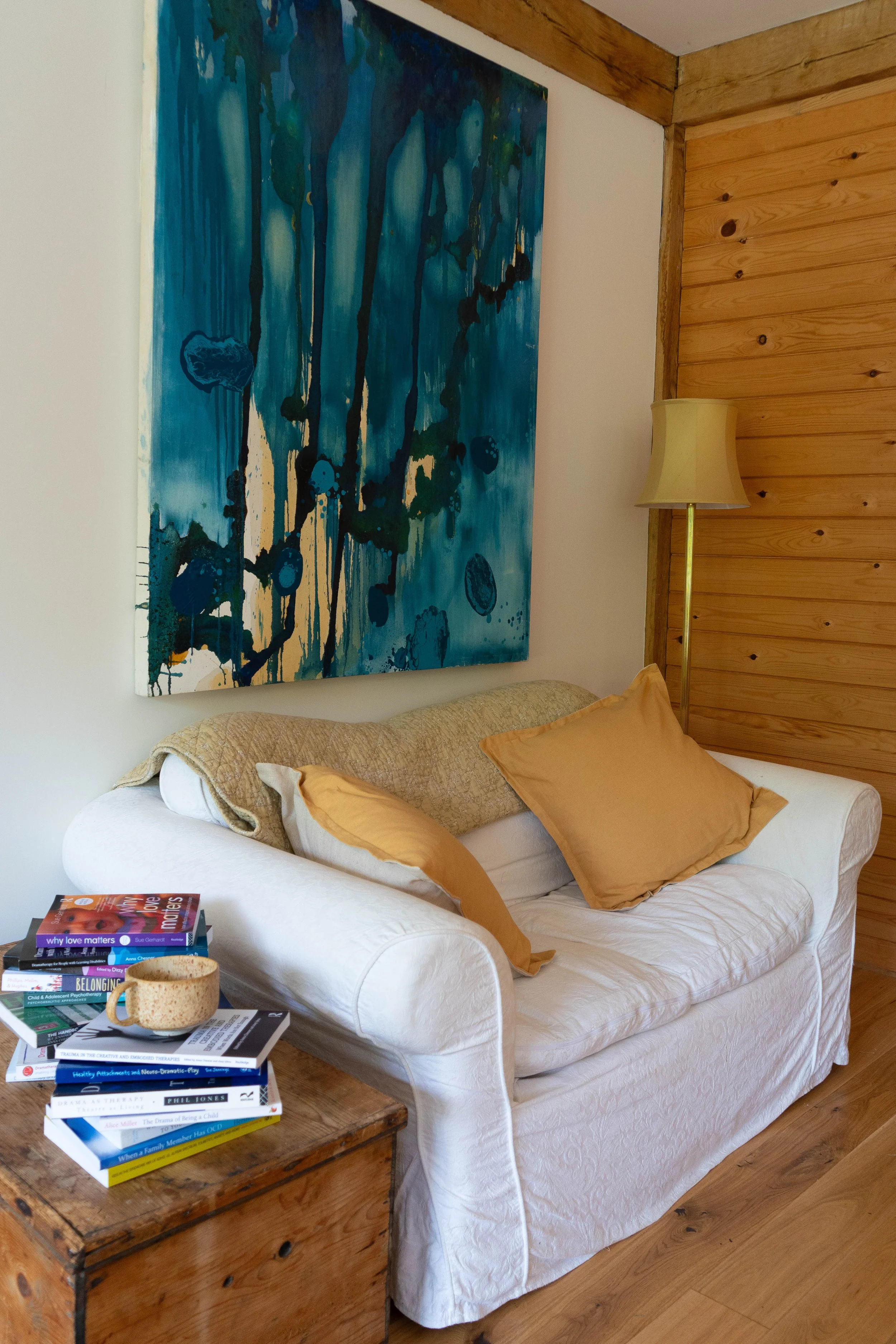 A cozy living room corner featuring a white upholstered armchair with a textured beige blanket and three mustard-colored pillows. Next to the chair is a rustic wooden side table stacked with books and a speckled ceramic mug. On the wall behind, there is a large, abstract blue painting and to the right, a wooden wall panel with a brass floor lamp.