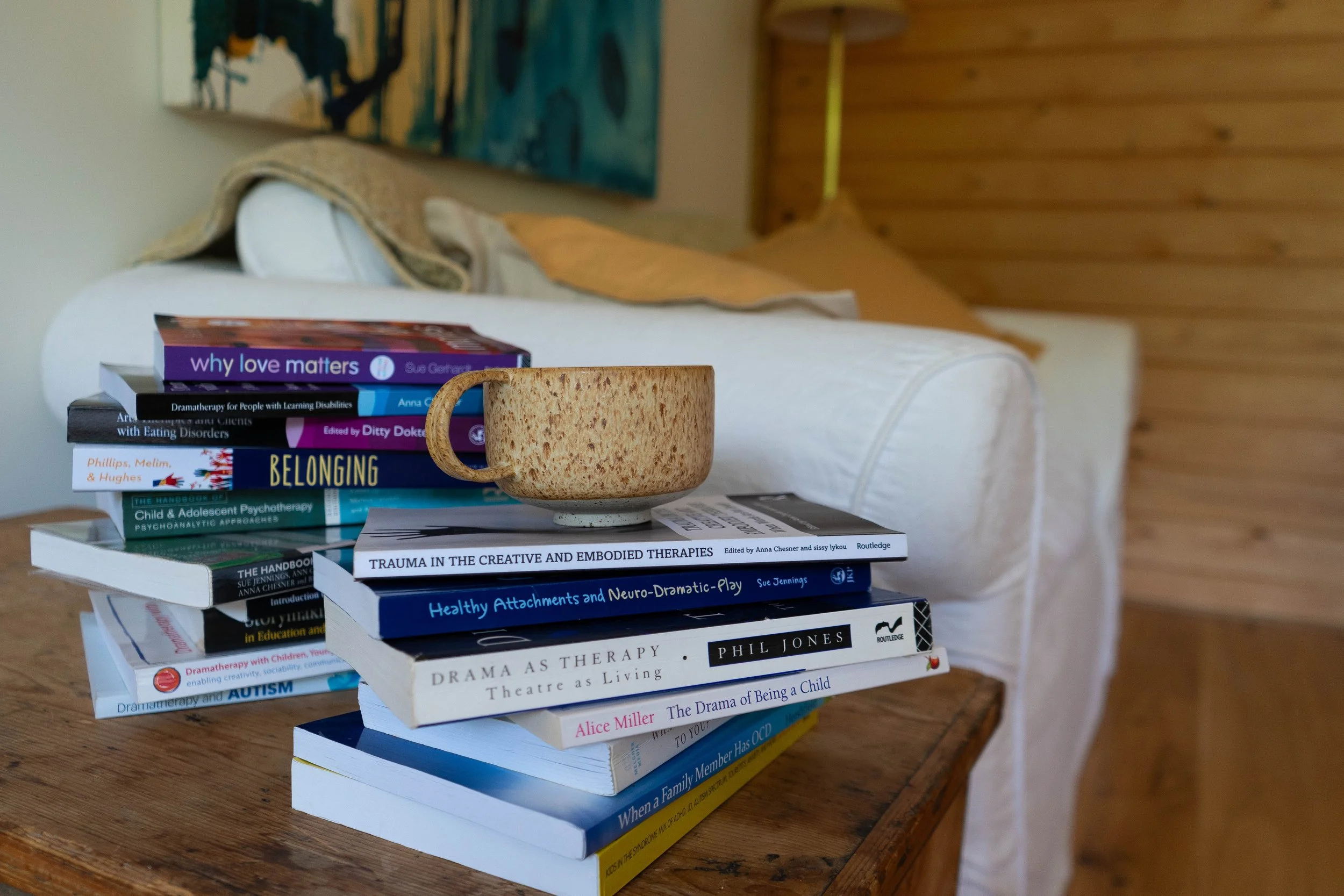 A stack of books on a wooden table with a beige mug in front of them. The books cover topics including love, learning disabilities, psychotherapy, attachment, trauma, autism, and childhood development. In the background, there is a white couch and a wooden wall, with a colorful abstract painting hanging behind the couch.