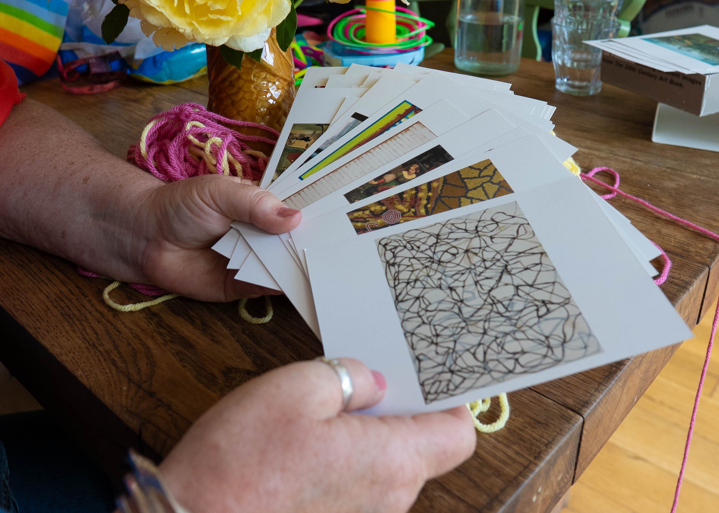 A person holding a fan of postcards with artwork on a wooden table, surrounded by colorful yarn, a flower arrangement in a vase, a candle, and various craft supplies.