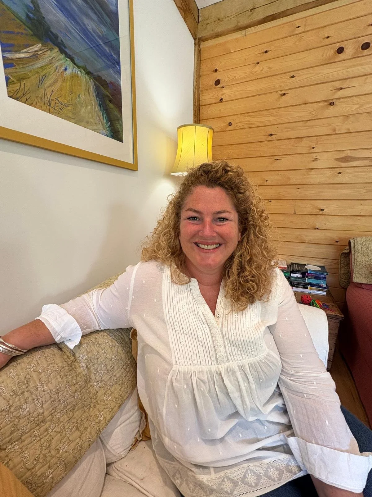 A woman with curly blond hair smiling while sitting on a beige patterned couch in a cozy living room with wooden walls, a yellow lamp, and a stack of books and DVDs on a side table.