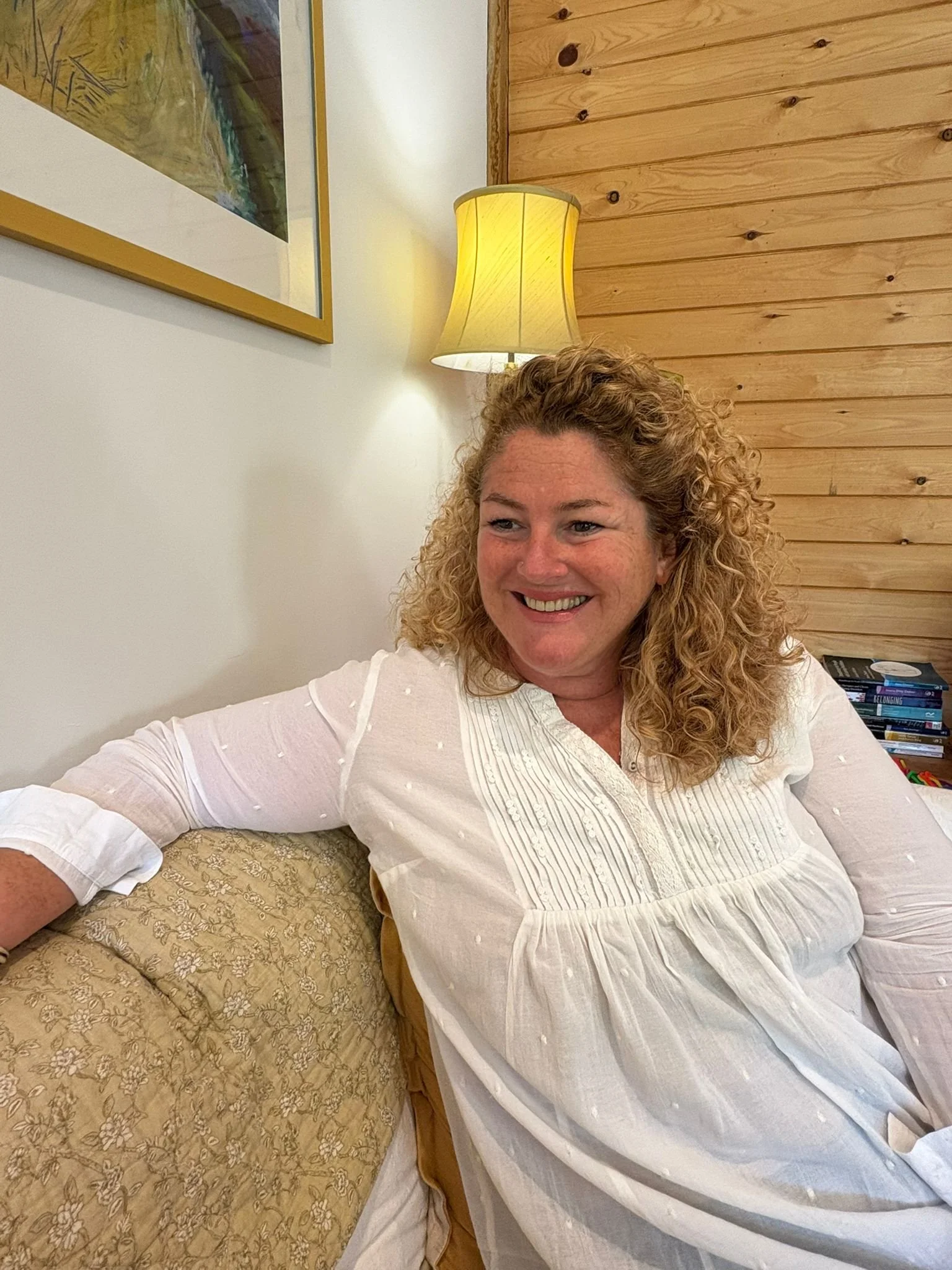 A woman with curly hair smiling, wearing a white blouse, sitting on a patterned beige sofa with a beige pillow. There is a wooden wall and a yellow lampshade in the background.