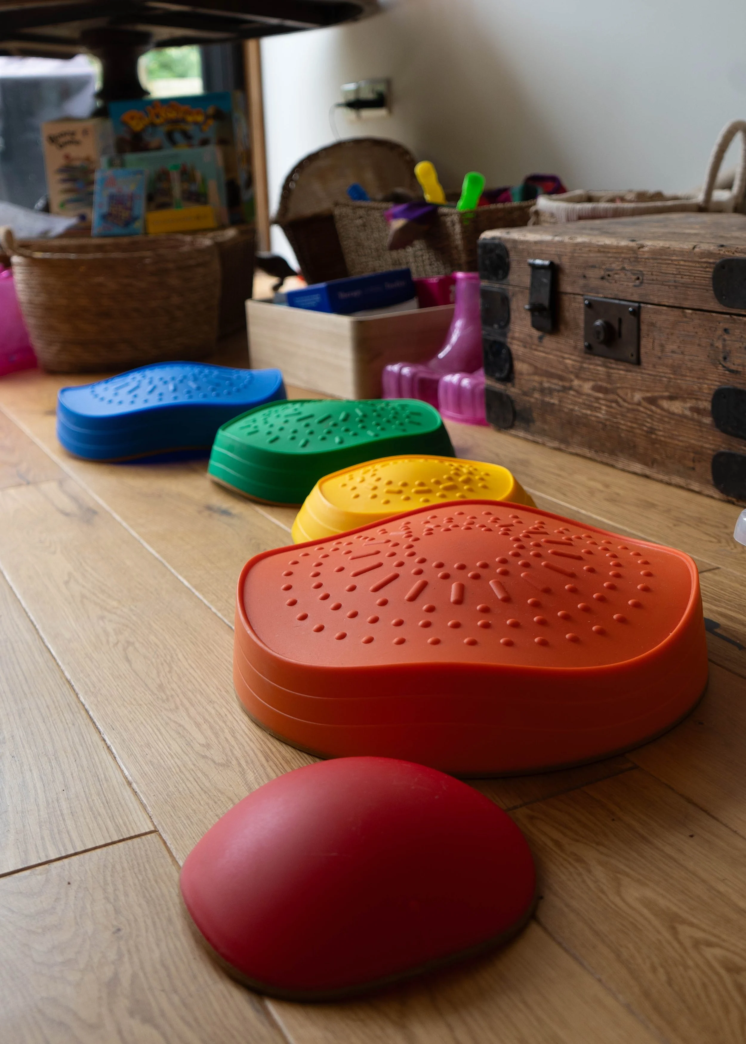 Colorful stepping stones in red, orange, yellow, green, blue, and purple on a wooden floor, with baskets and toys in the background.