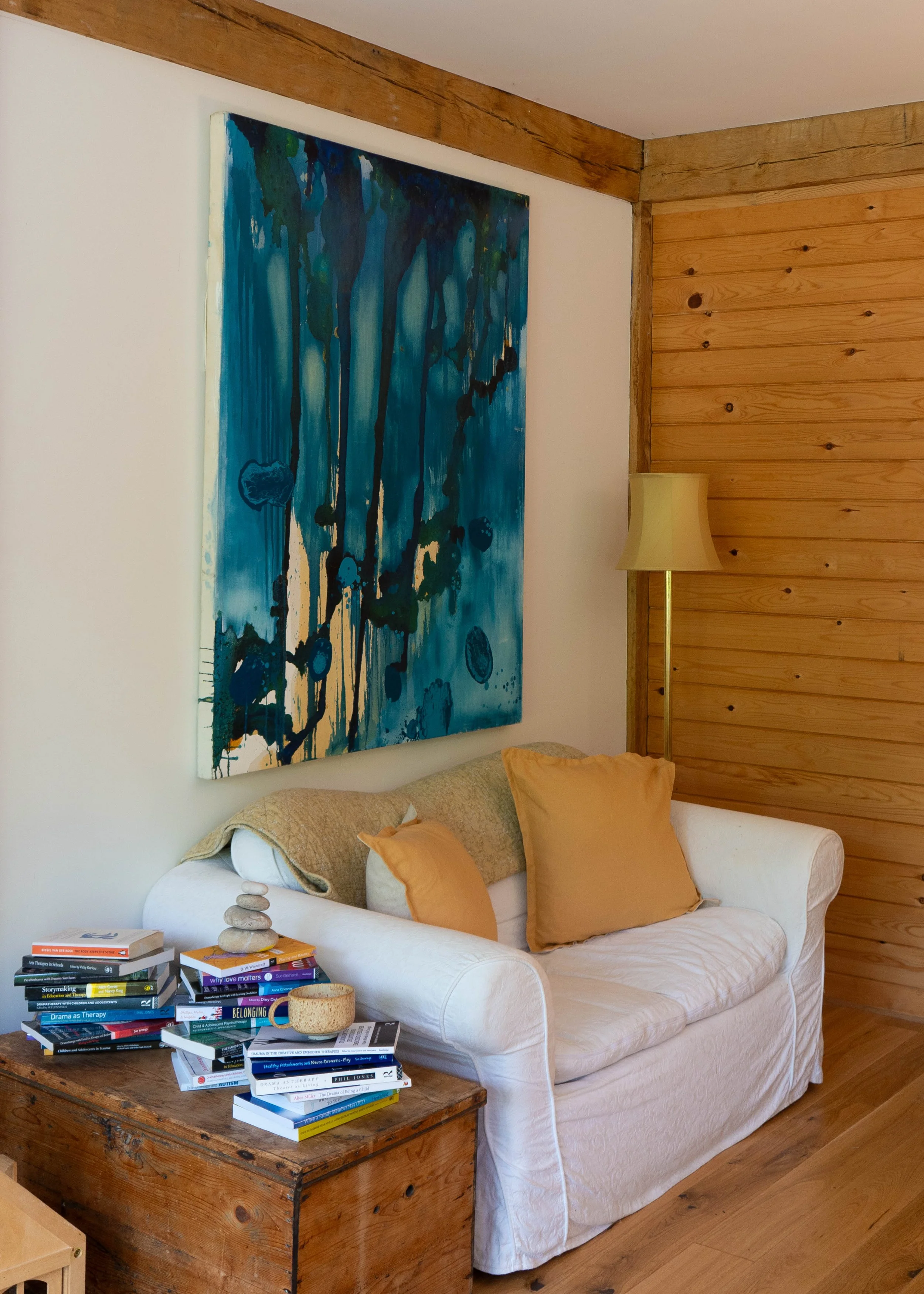 A cozy living room corner with a white sofa, tan cushions, and a large abstract blue and green painting on the wall. There is a wooden chest used as a coffee table with a stack of books and a small bowl, and a yellow floor lamp next to the sofa. The walls are a mix of white paint and wood paneling.