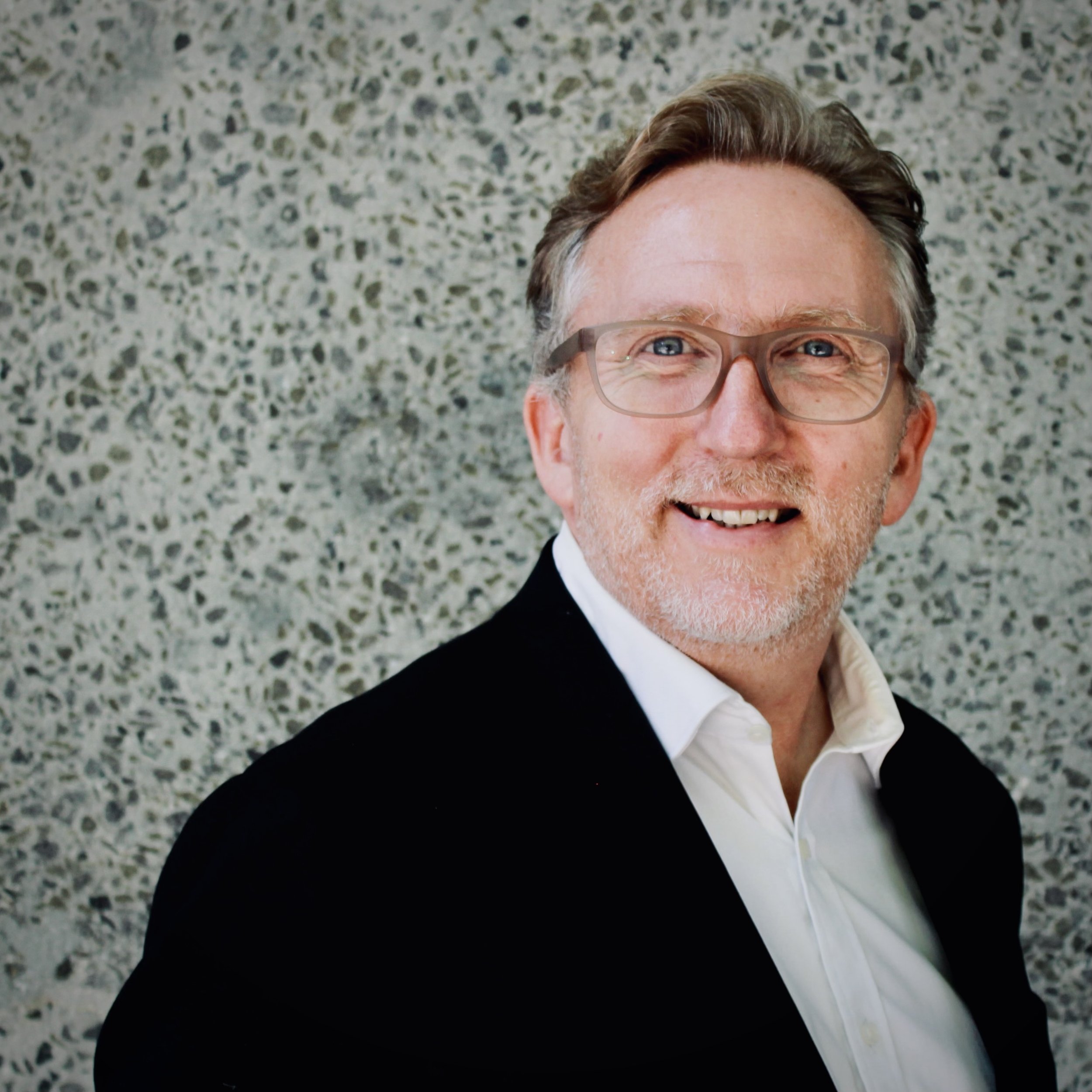 A smiling middle-aged man with glasses, wearing a tuxedo and a bow tie, is looking at the camera while standing on a speckled floor.