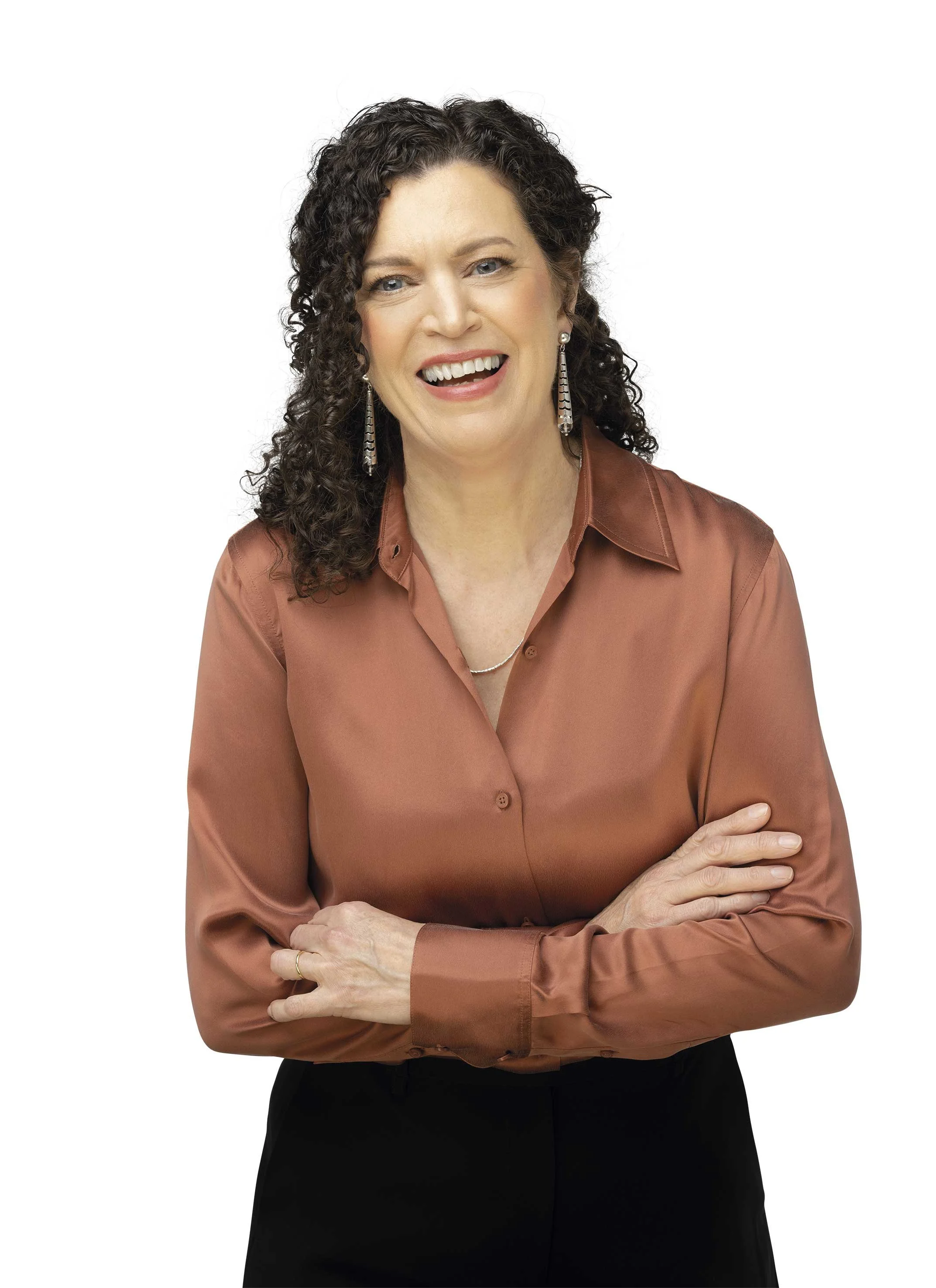 A woman with curly brown hair wearing a copper-colored blouse and black pants. She is smiling with her mouth open and has her arms crossed.