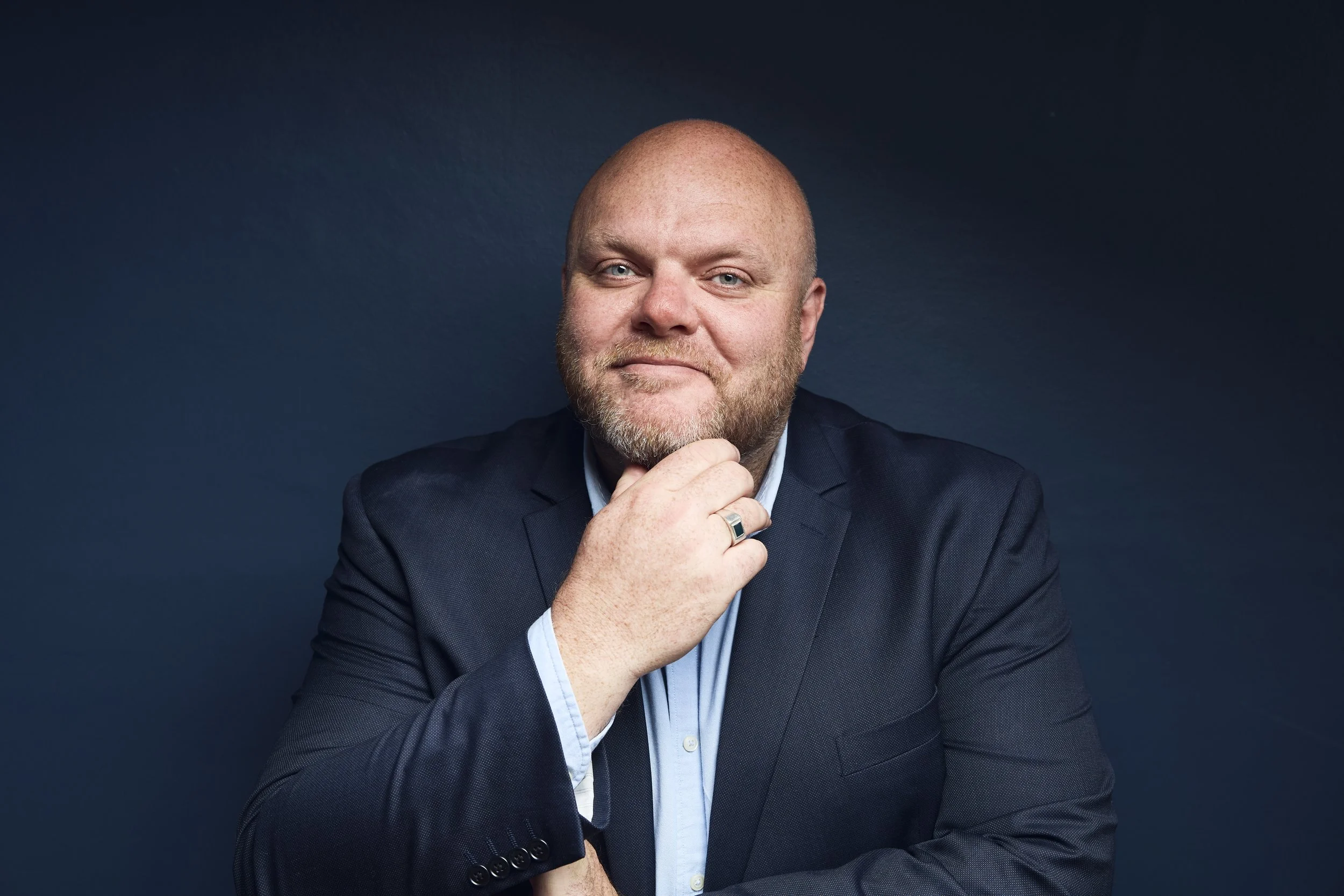 A bald man with a beard in a navy suit and light blue shirt, resting his chin on his hand, looking at the camera against a dark blue background.