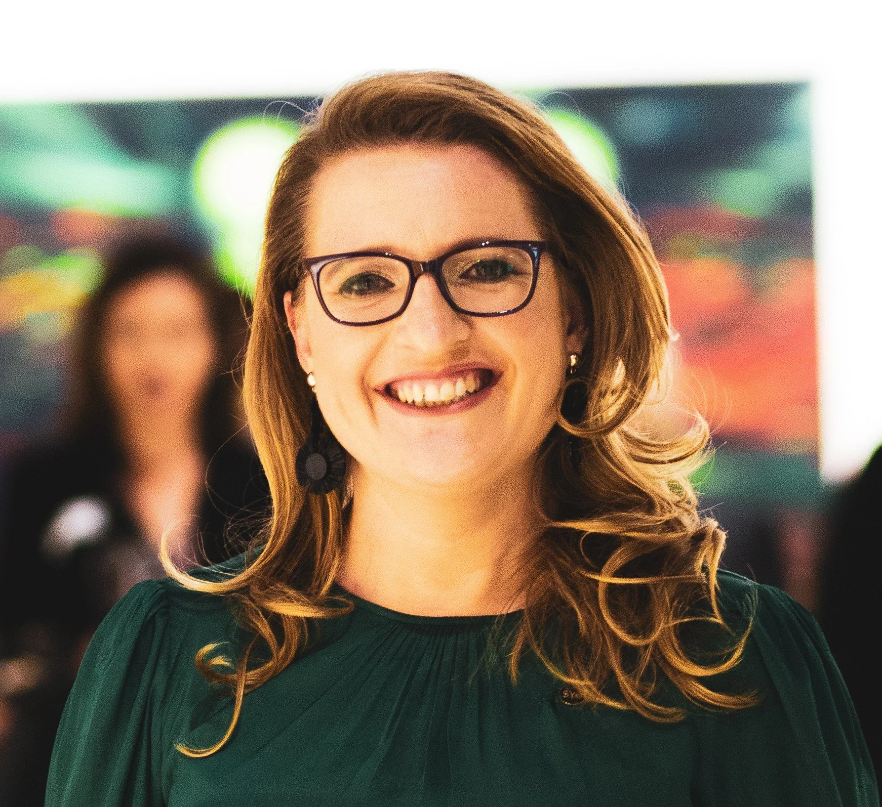 A woman with long, wavy red hair, wearing glasses and a green dress, smiling in an indoor setting with colorful lights in the background.