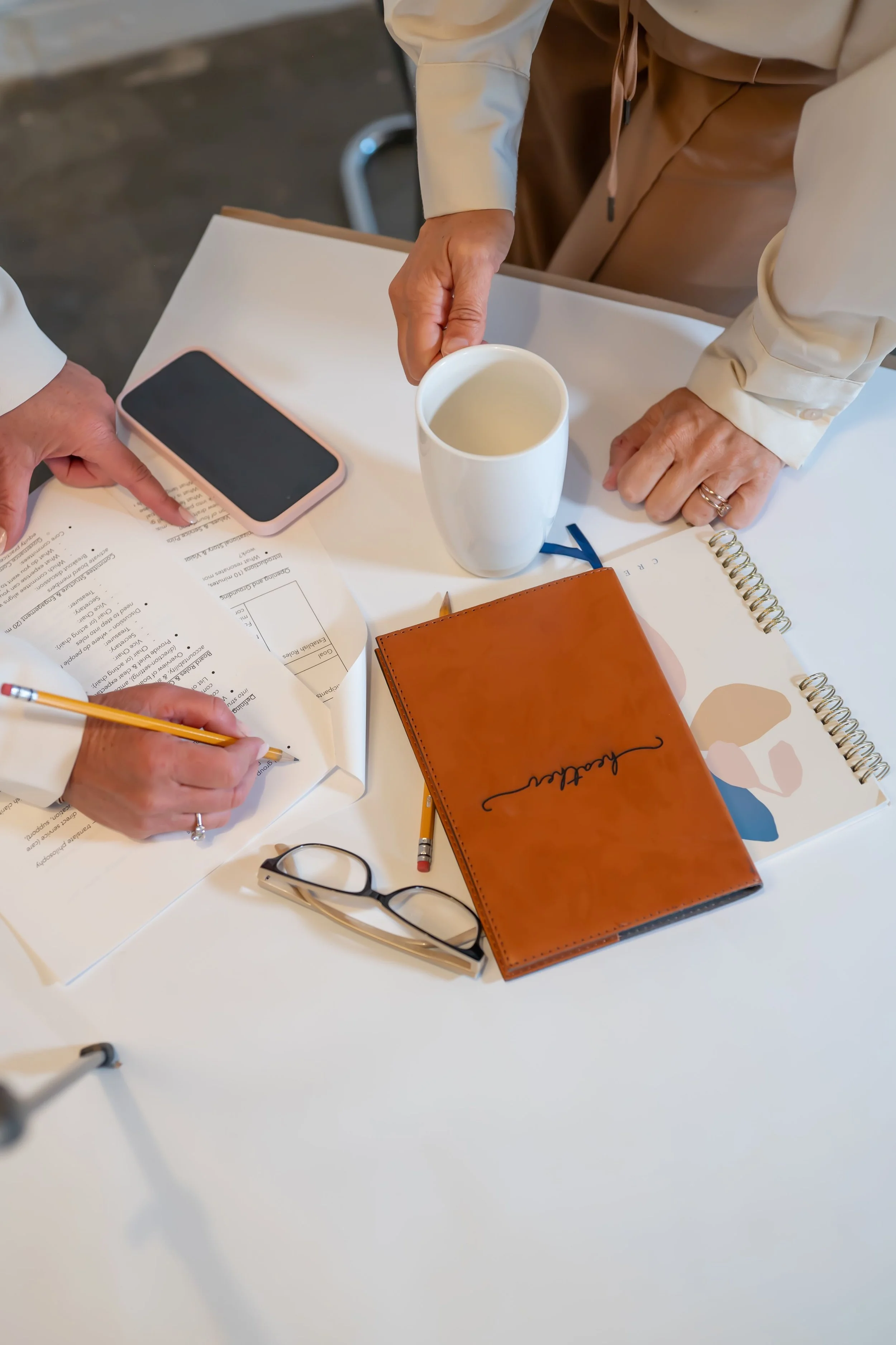 Flat lay of table with a woman writing on a checklist and another woman grabbing for coffee mug next to phone and notebooks on table